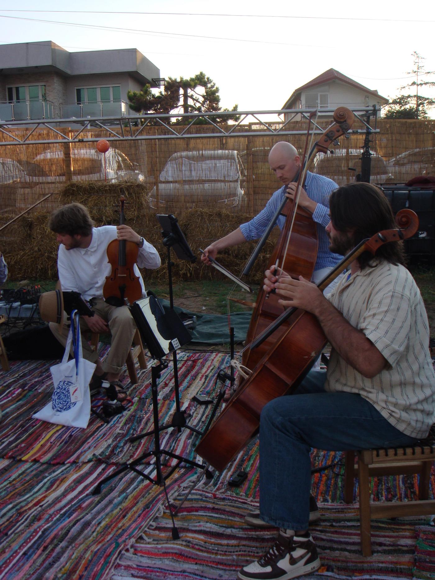 Two musicians in soundcheck on a rag rug laid over sand, hay bales and a lighting truss behind them in low evening sun.