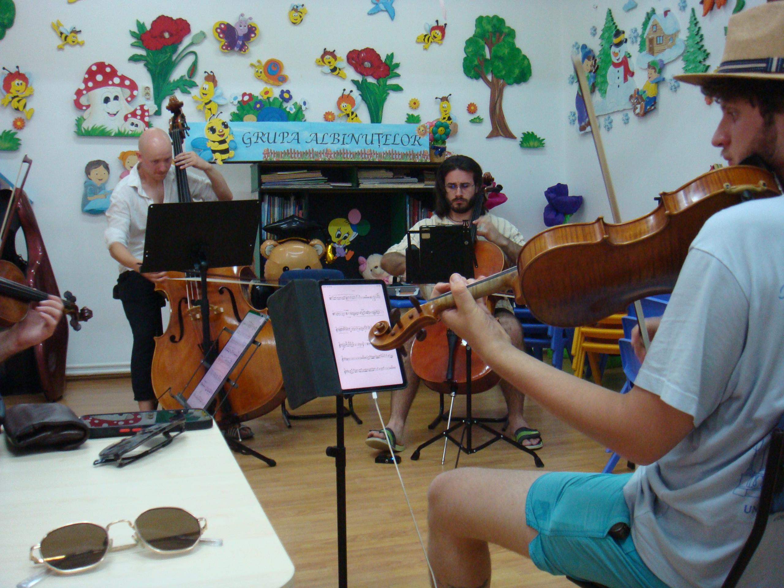 Three musicians rehearsing in a kindergarten classroom, a wall of cartoon bees and flowers behind them, a violin scroll in the foreground.