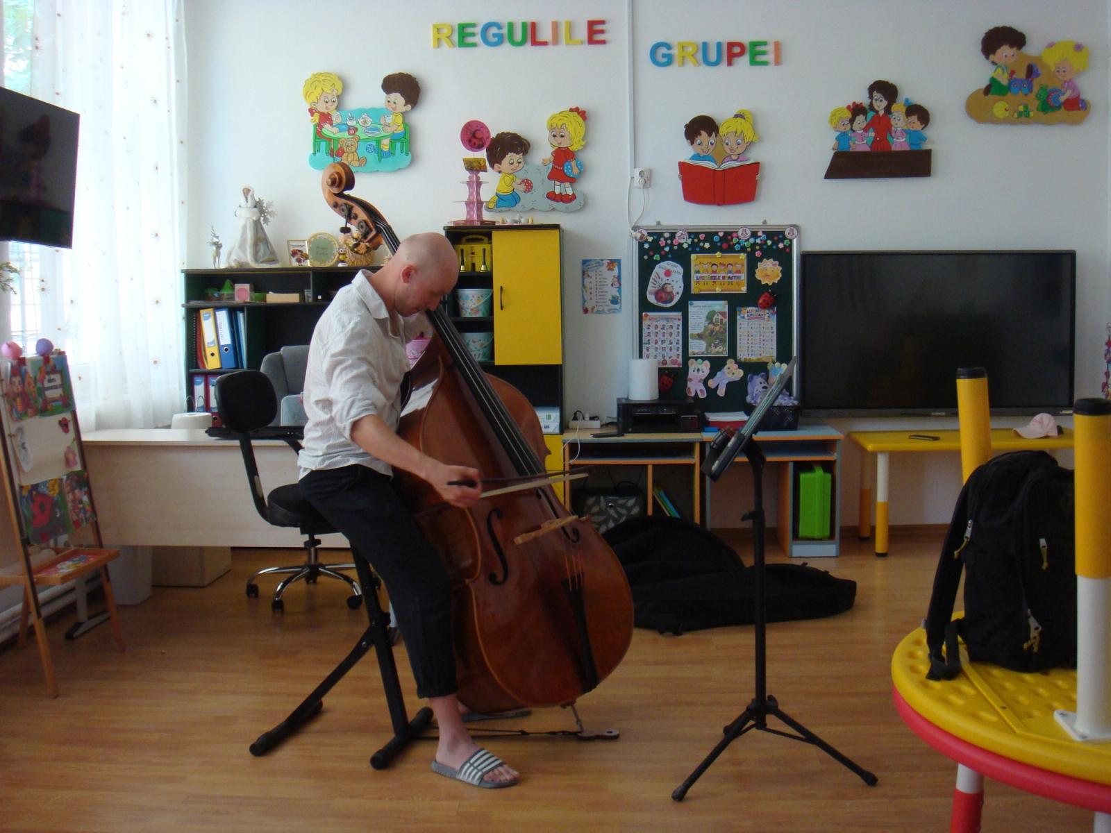 A double bass player alone in a kindergarten classroom, bowing, beneath a wall display headed REGULILE GRUPEI, the rules of the group.