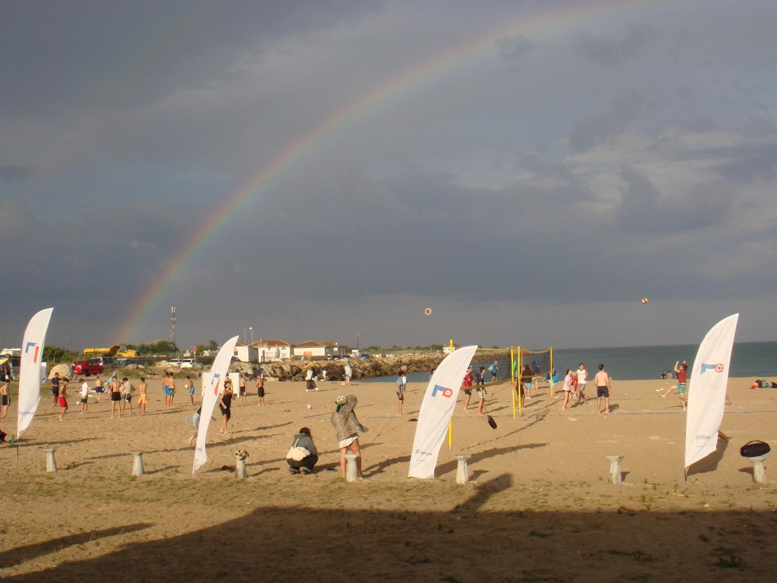A rainbow over the beach at 2 Mai, festival flags in the sand, people playing volleyball under a dark sky.