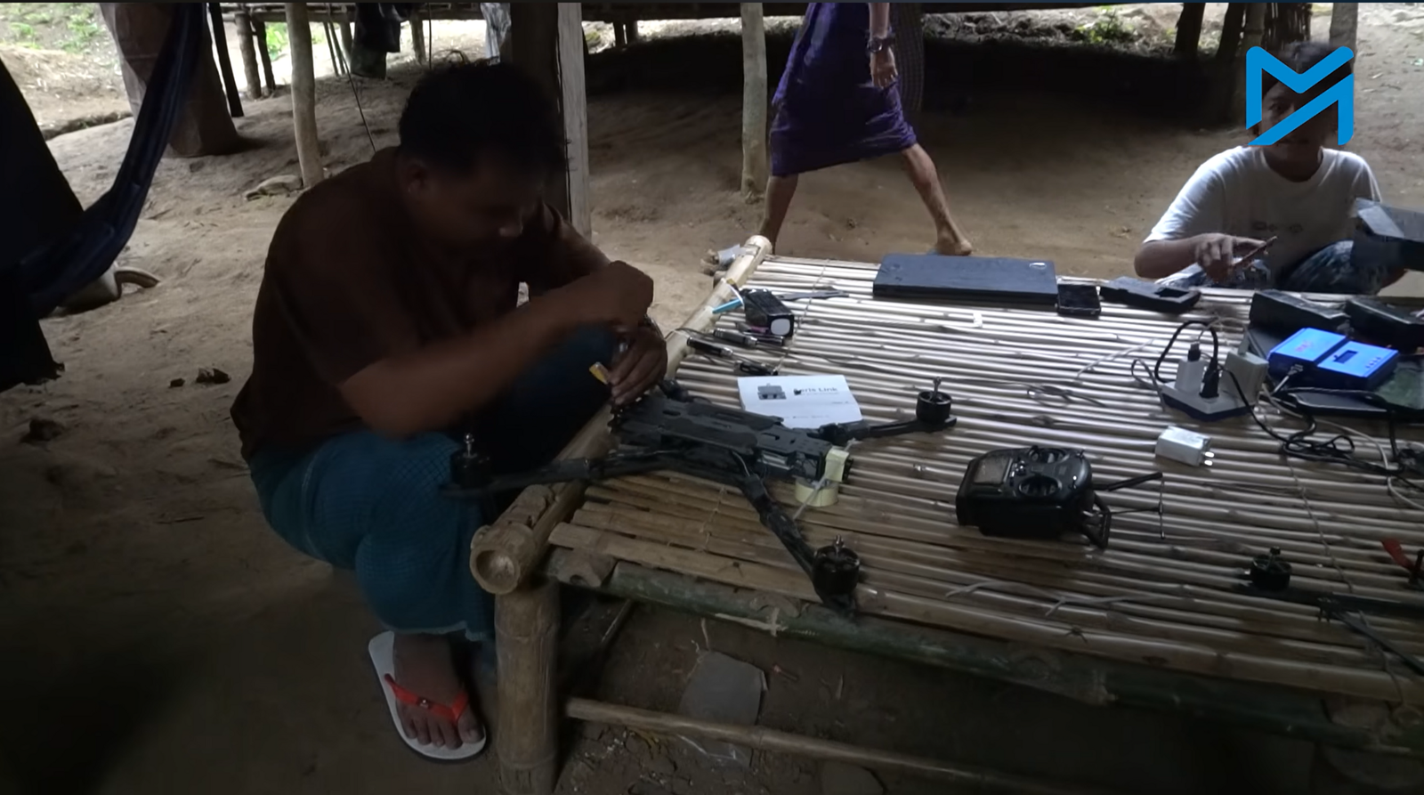 A man in sandals kneels by a low table as he assembles a small drone.