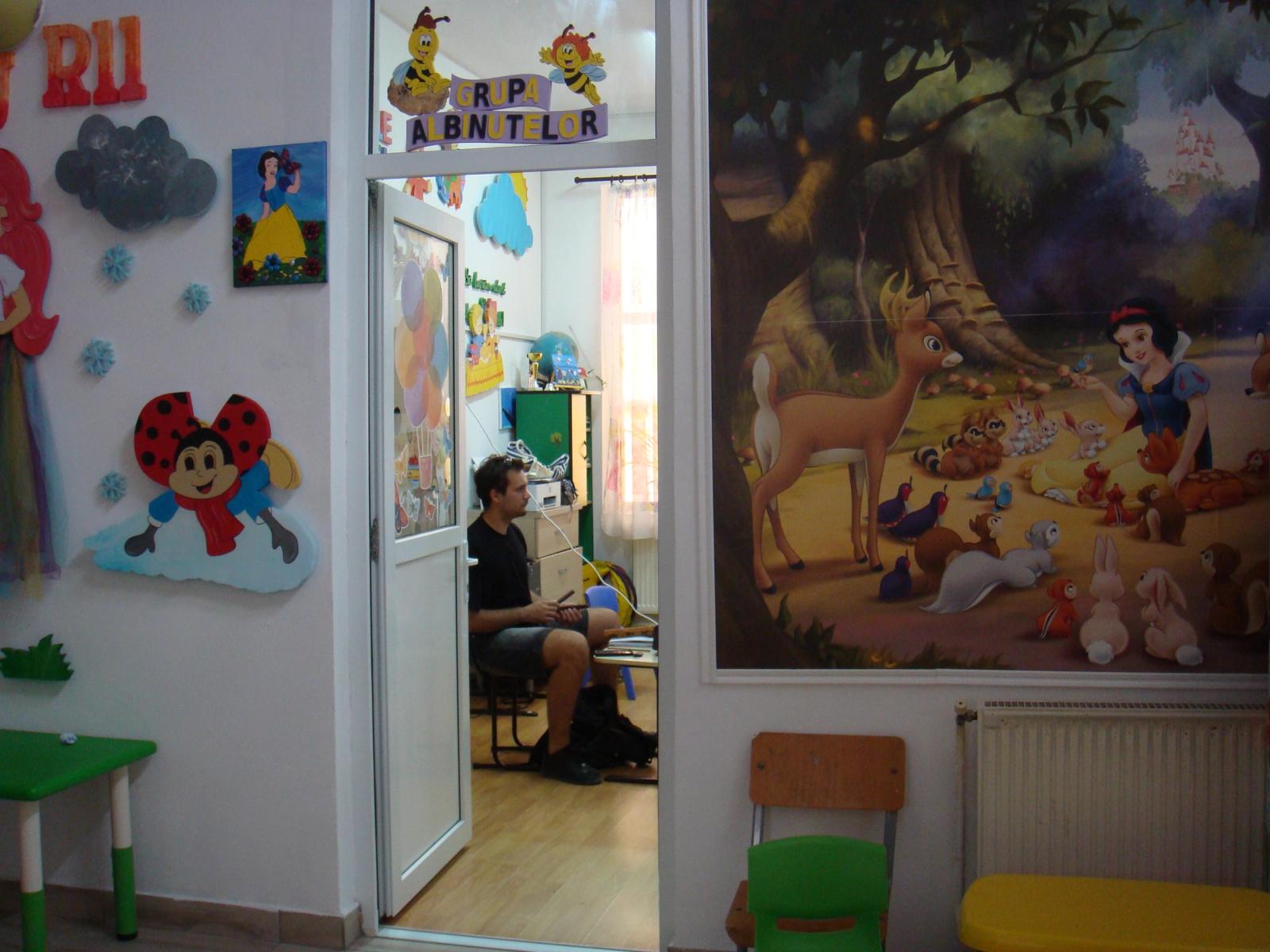 A kindergarten corridor with a Snow White mural and a ladybird cutout, a musician visible through an open door, sitting alone with his bow.