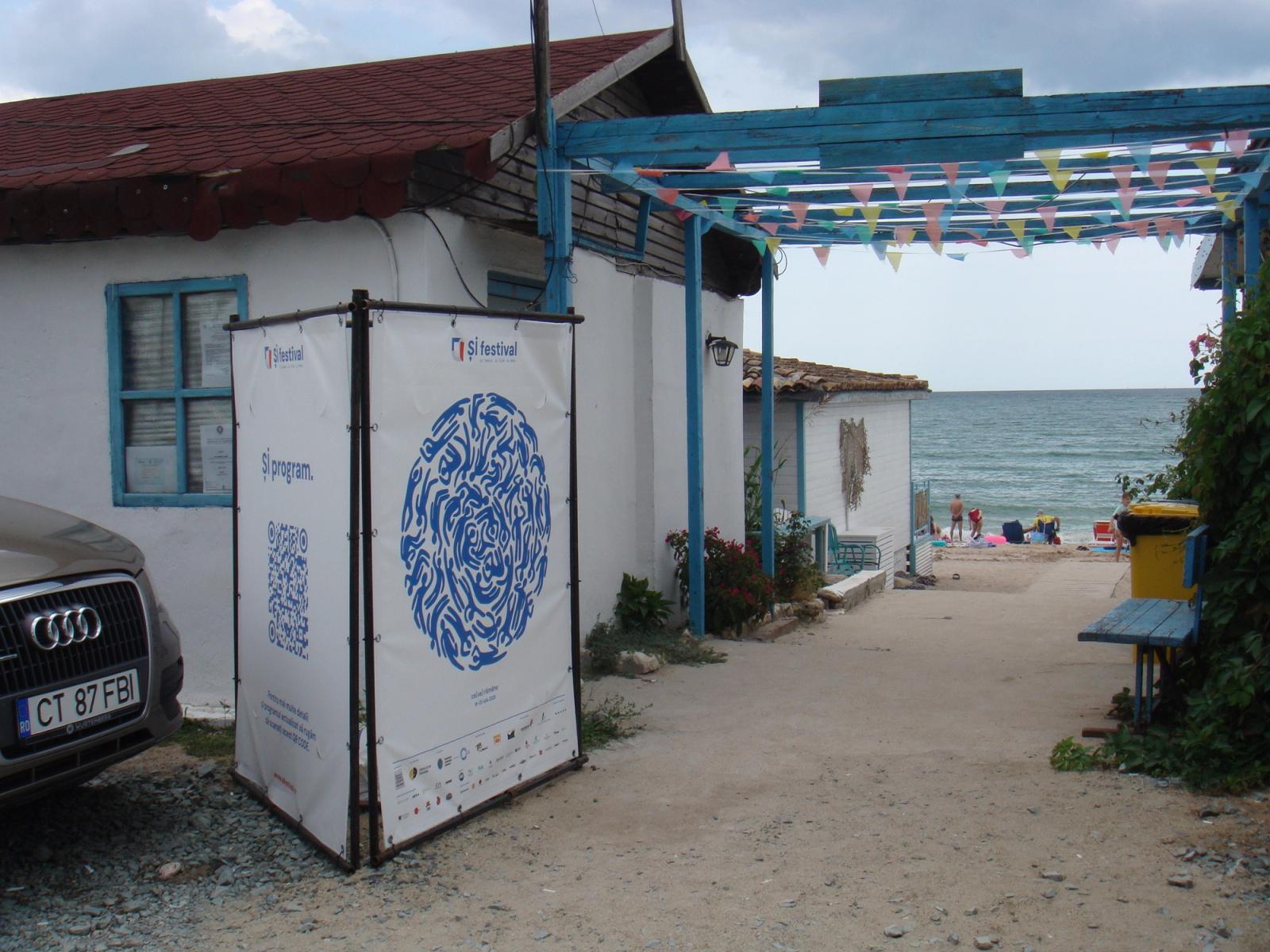 A ȘI festival programme banner outside a whitewashed seaside building, bunting overhead and the Black Sea beyond.