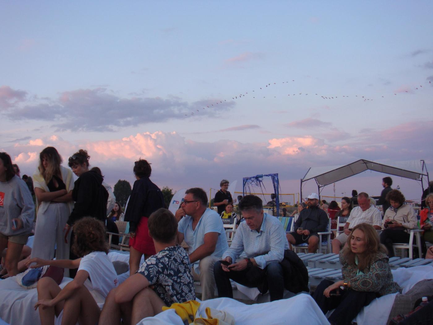 The festival audience settling onto white loungers on the sand at dusk, pink cloud above them and a line of birds crossing the sky.