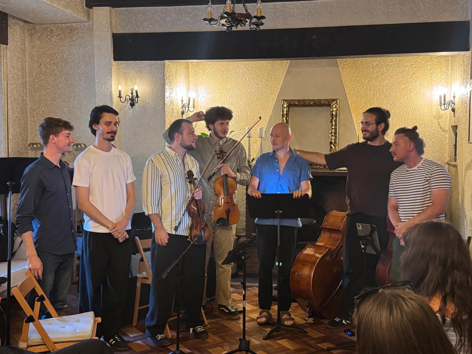 Seven young men standing together in a living room after playing, holding a violin and a viola, a double bass behind them.