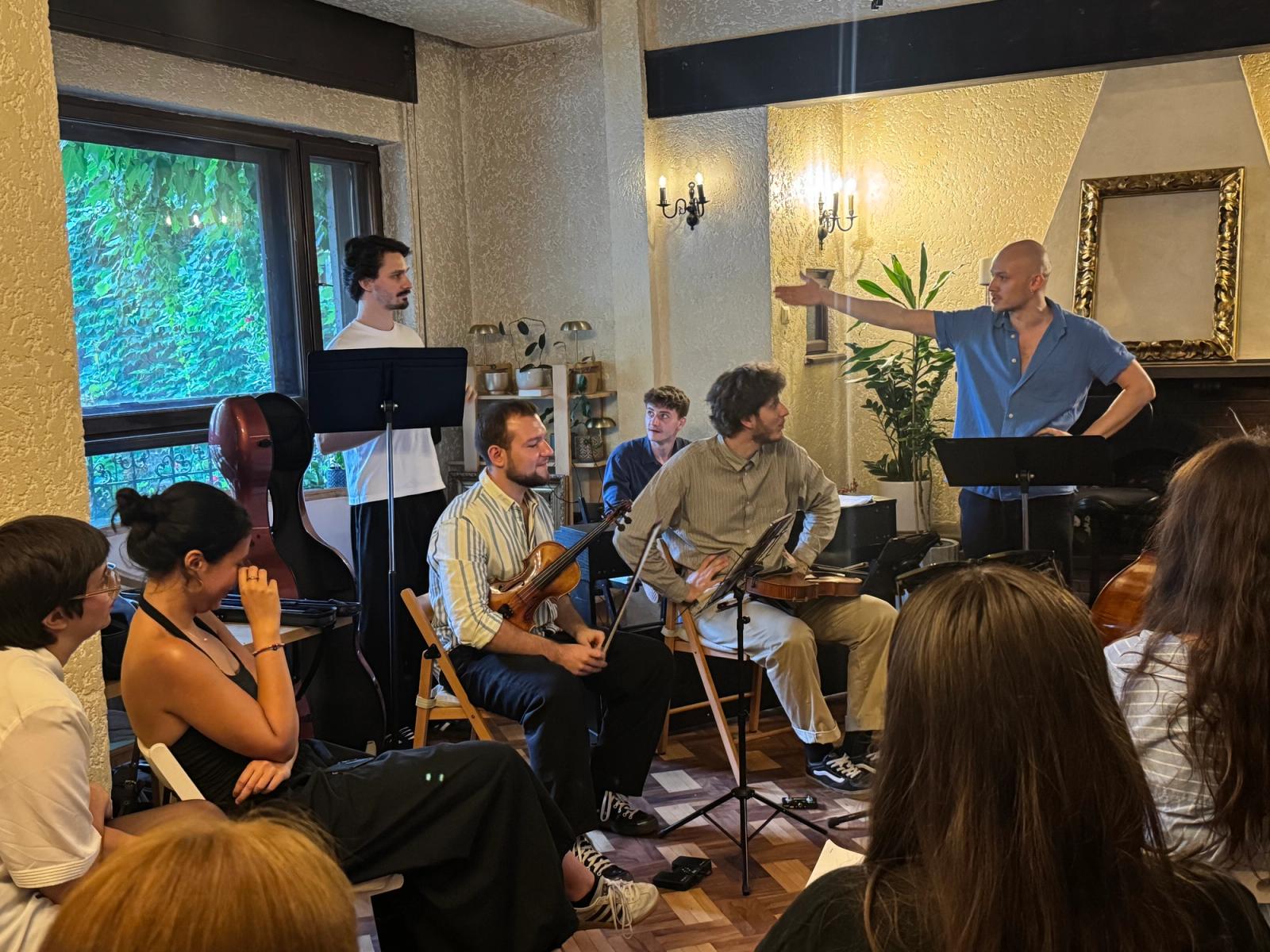 A standing musician gestures mid-sentence while four others sit with their instruments, an audience seated close around them.