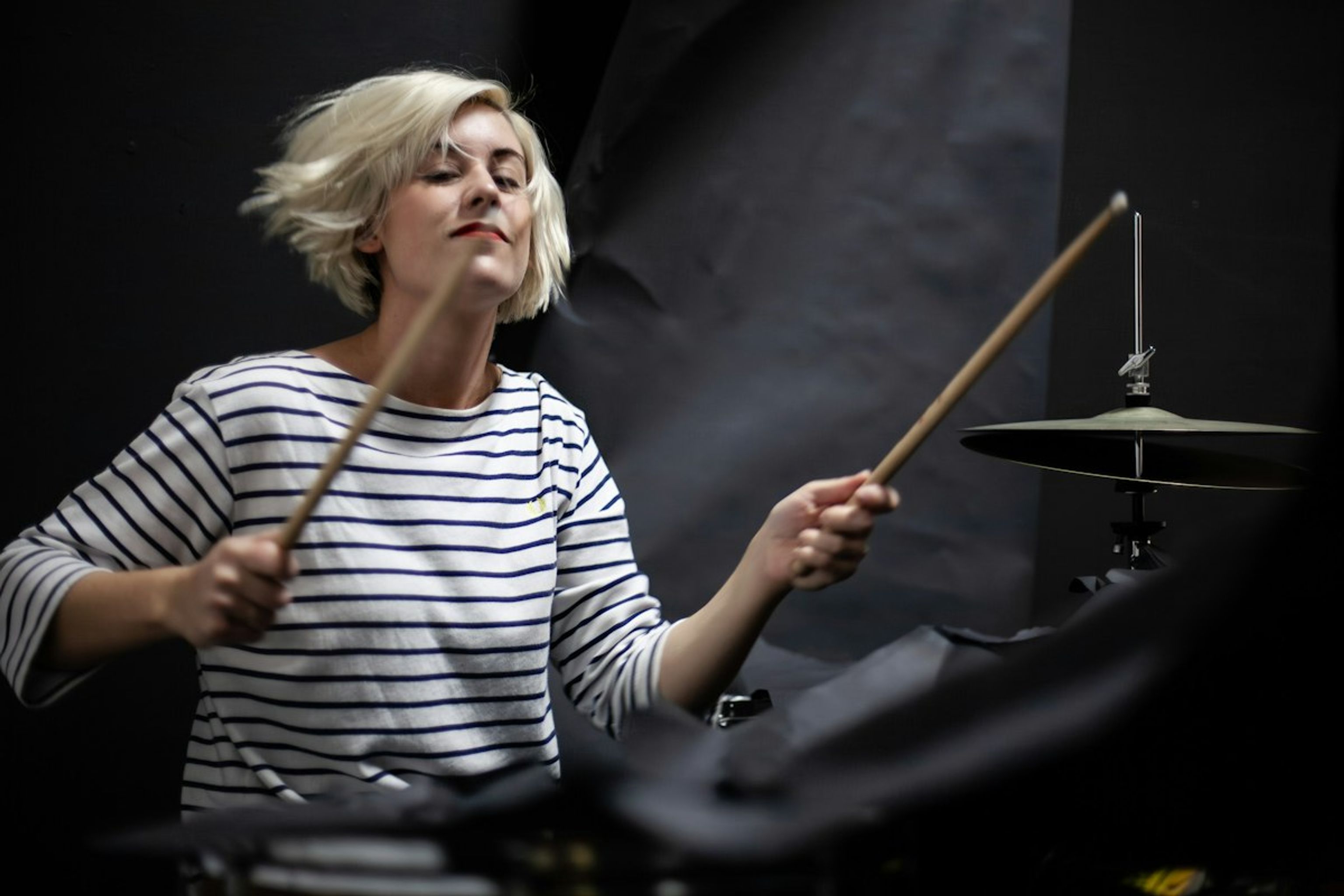 Woman with short blonde hair in a striped top playing the drums, eyes closed