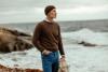 A man in a brown crew-neck sweater and beanie on a rocky shoreline