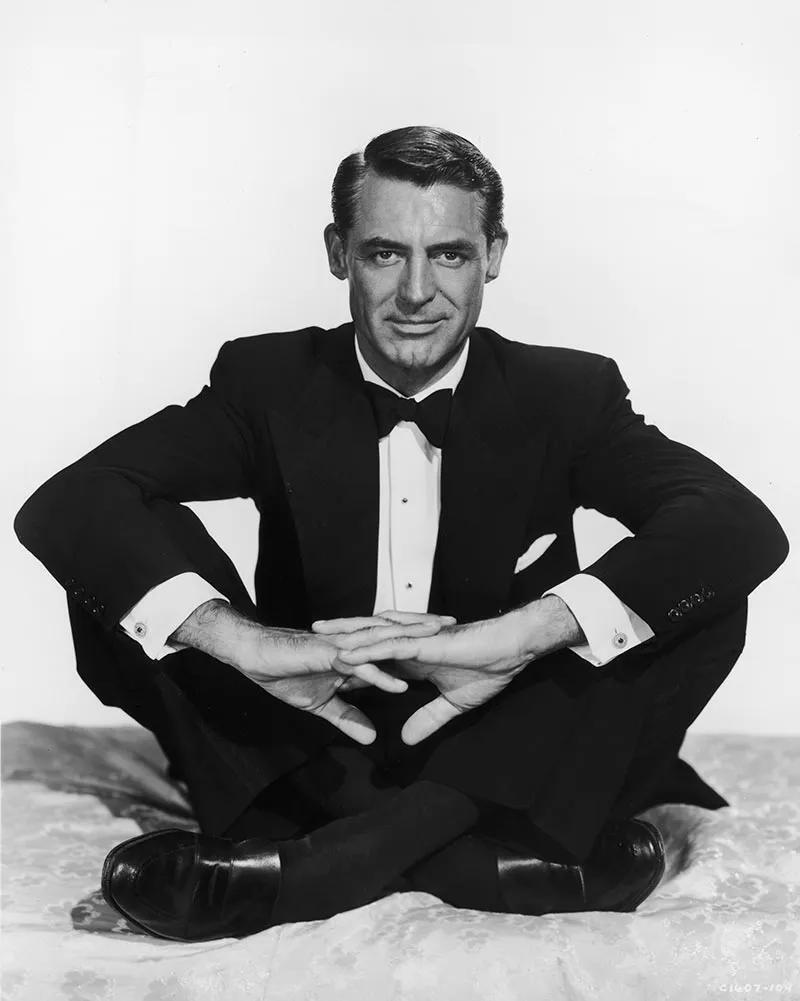 Cary Grant in a black tuxedo and bow tie, seated cross-legged on the floor