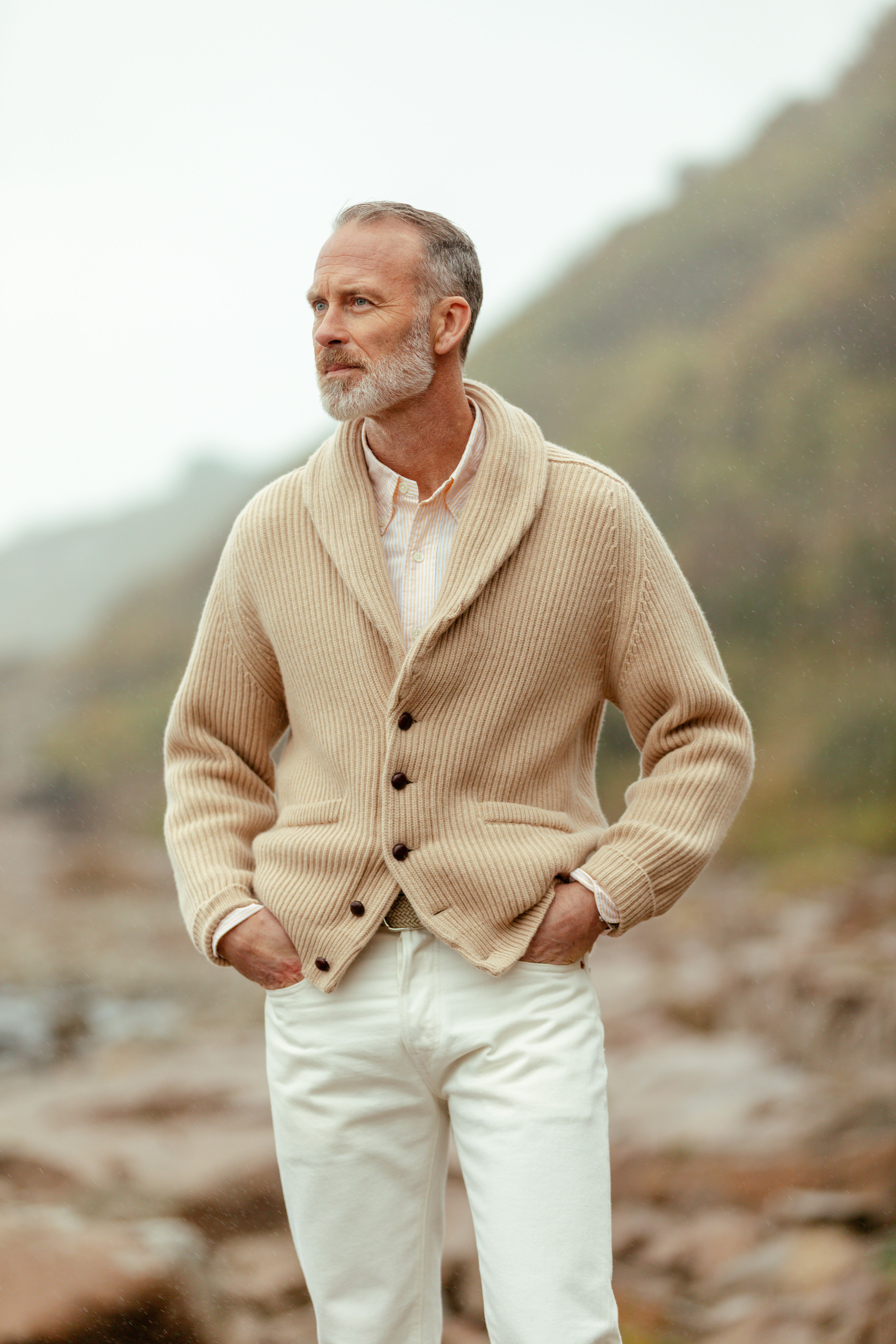 A man in a cream shawl-collar cardigan and white trousers in a hillside valley