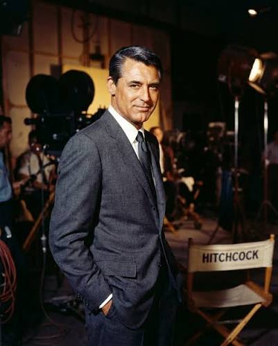 Cary Grant in a grey suit on a film set, beside a director's chair marked Hitchcock
