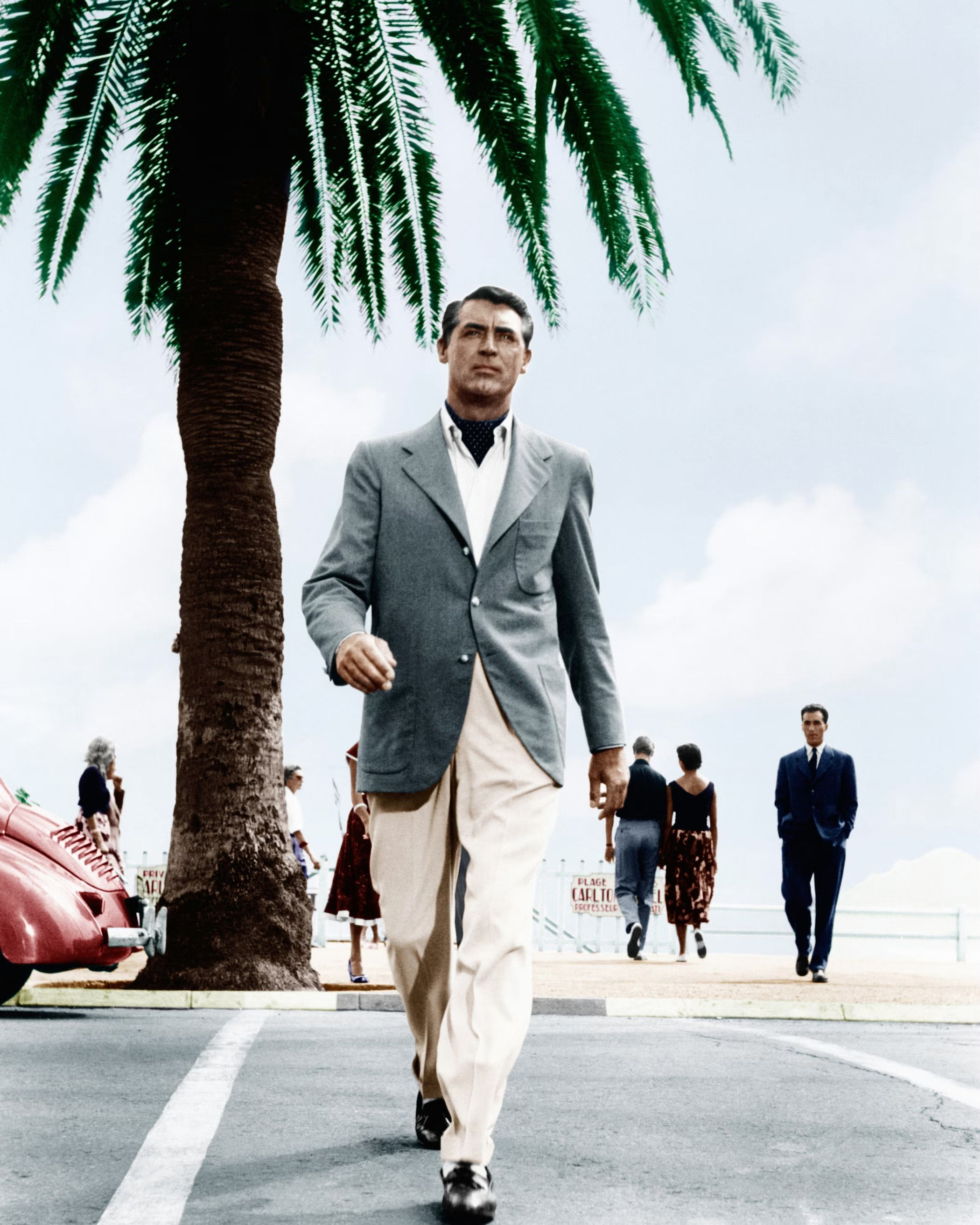 A man in a light jacket and cream trousers walking along a palm-lined street