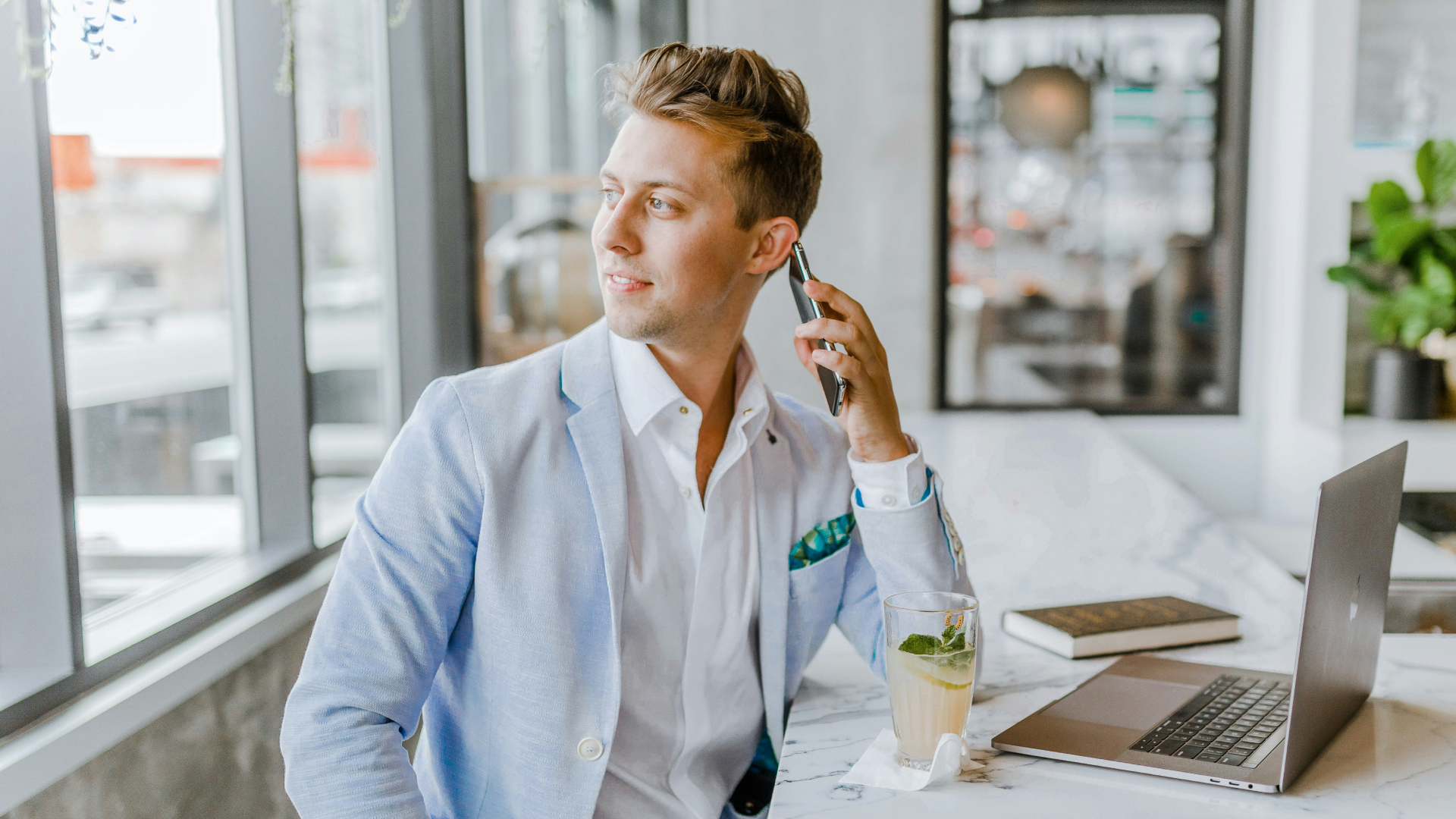 Man in a light blue blazer working on a laptop while speaking on his cell phone.