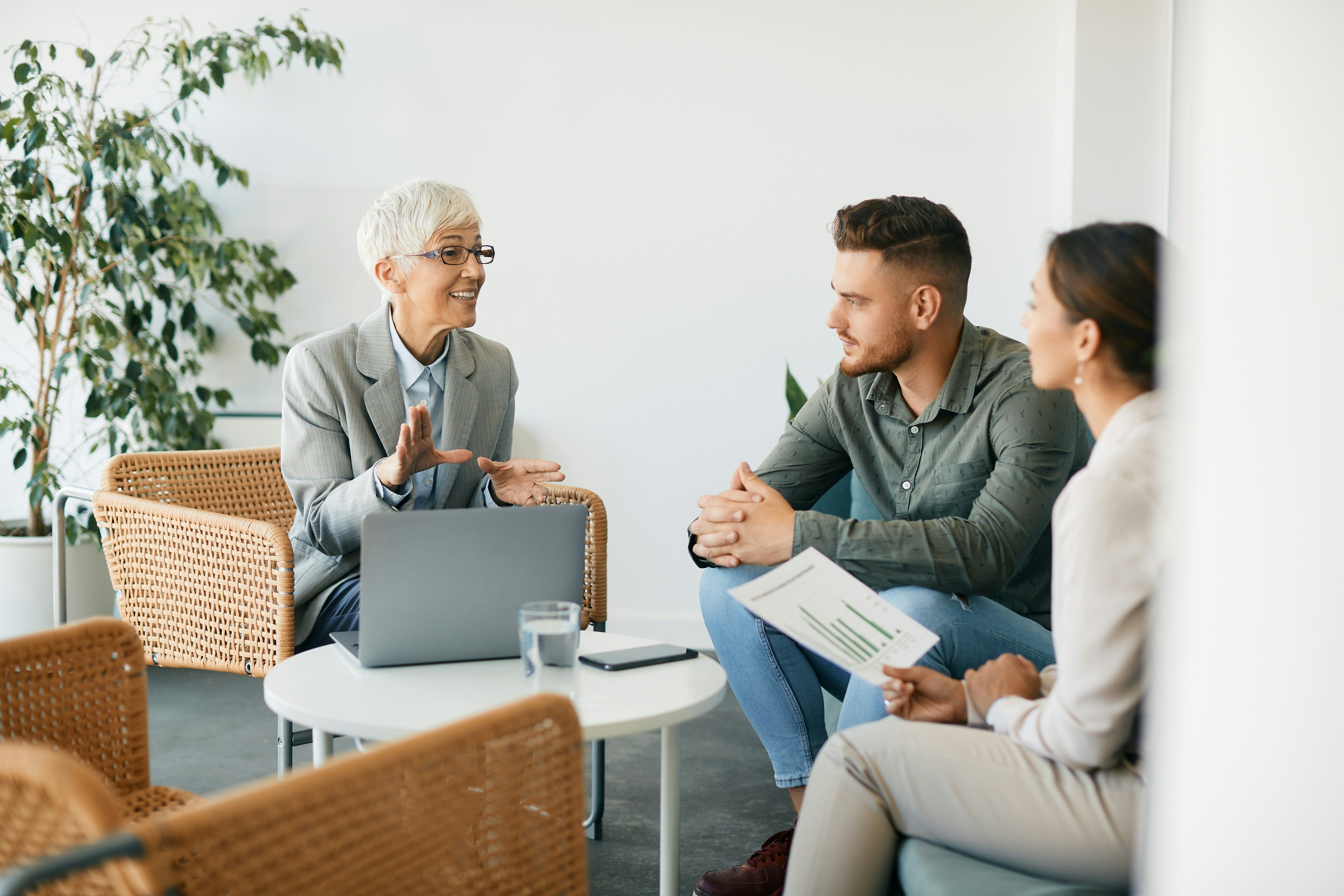 Woman with glasses and a grey suite talking to two colleagues over a coffee table with a laptop