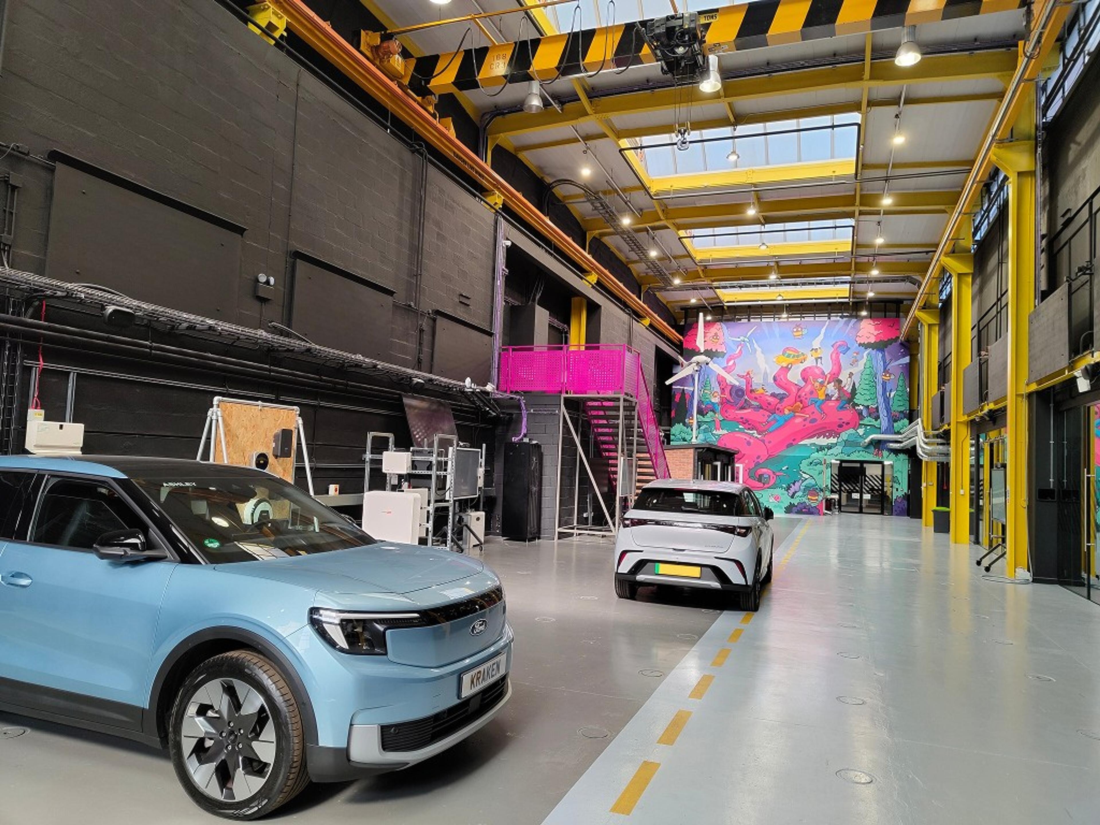 Interior of a large industrial-style hall with a light blue Ford electric SUV and a white BYD car on the floor, and a colourful mural at the far end.