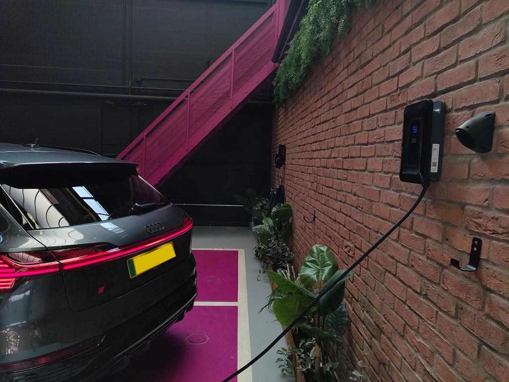 Dark grey Audi e-tron parked beside a wall-mounted home EV charger on an exposed brick wall, with plants and a magenta painted bay.