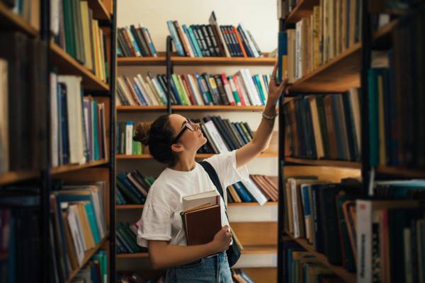 En student som holder pensumbøker i hånden og leter etter bøker på biblioteket.