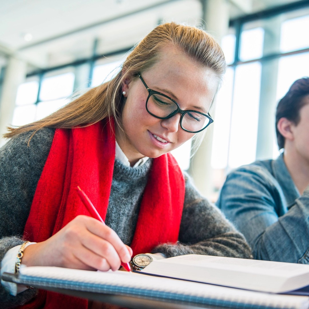 Illustrasjonsfoto. Studenter. I forgrunnen er en kvinnelig student, med en bok åpen på bordet foran seg, som skriver i en notatblokk.
