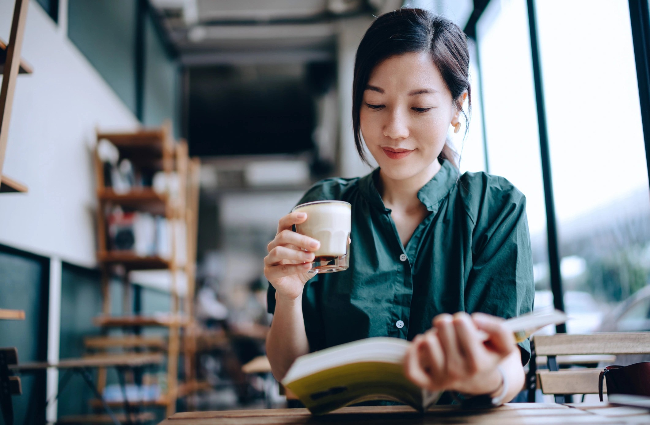 En ung kvinne sitter og leser en bok mens hun holder en kaffe i hånden.