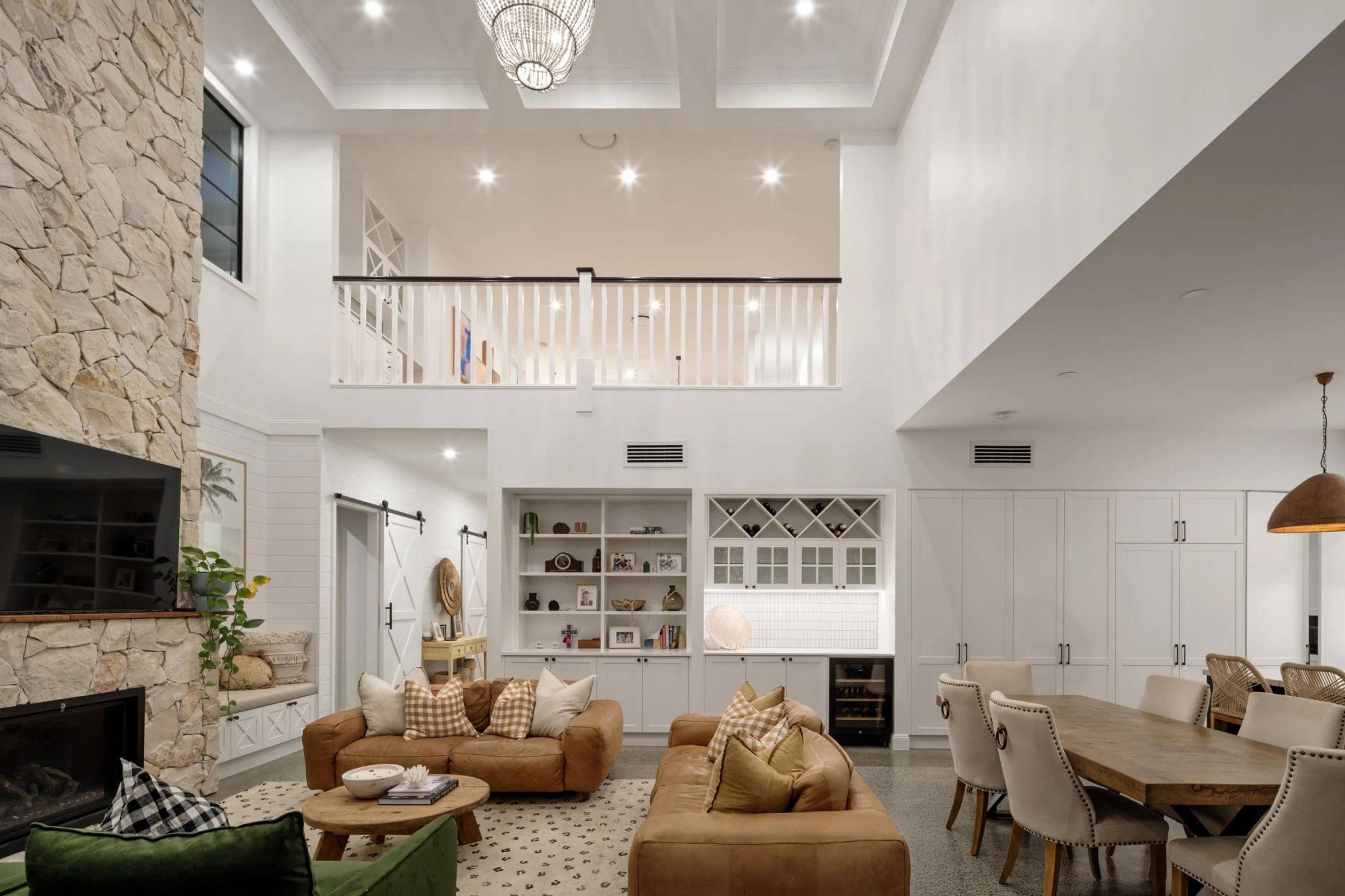 Luxury double-height open-plan living and dining room featuring a stone fireplace, custom cabinetry, and mezzanine level by Invilla Architecture