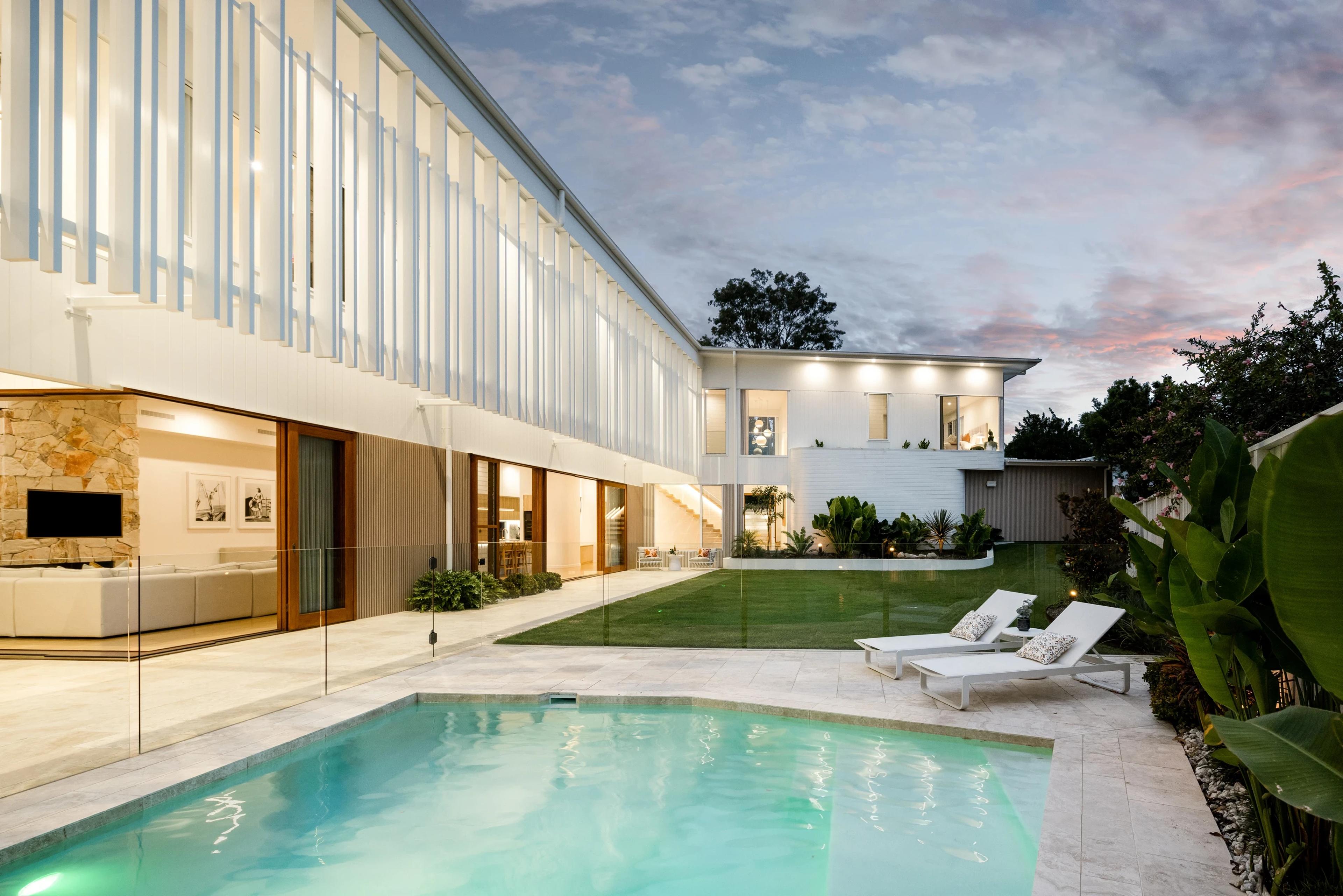 Poolside terrace and lawn beside a white two storey home at dusk