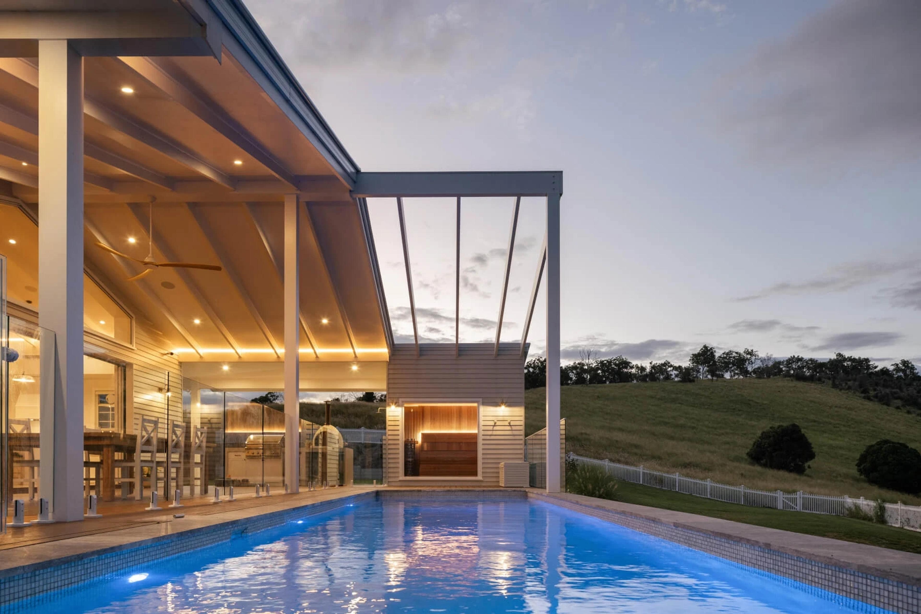 Swimming pool and lit verandah at dusk beside grassy hill