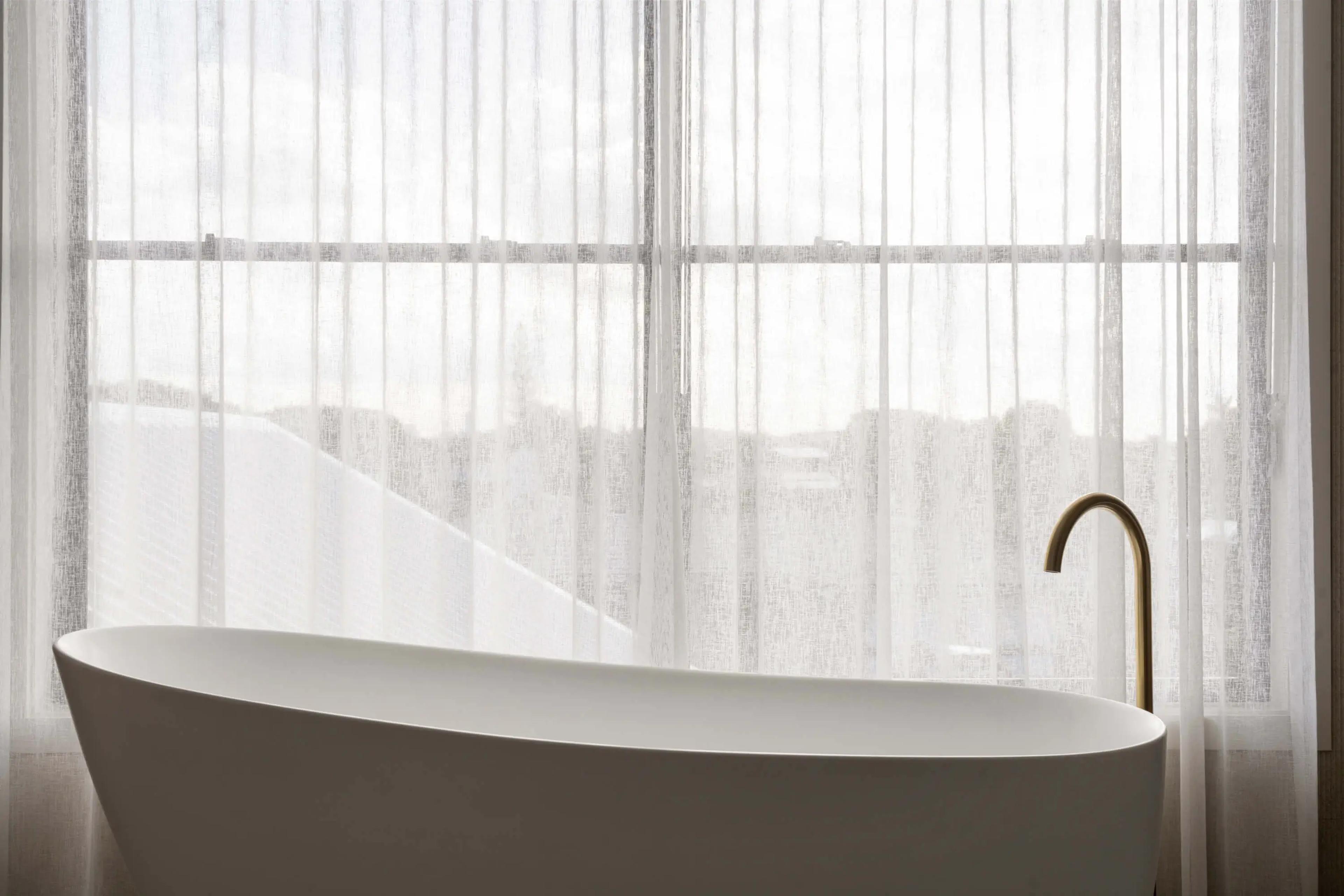 Freestanding bath by sheer curtained window with brass tap
