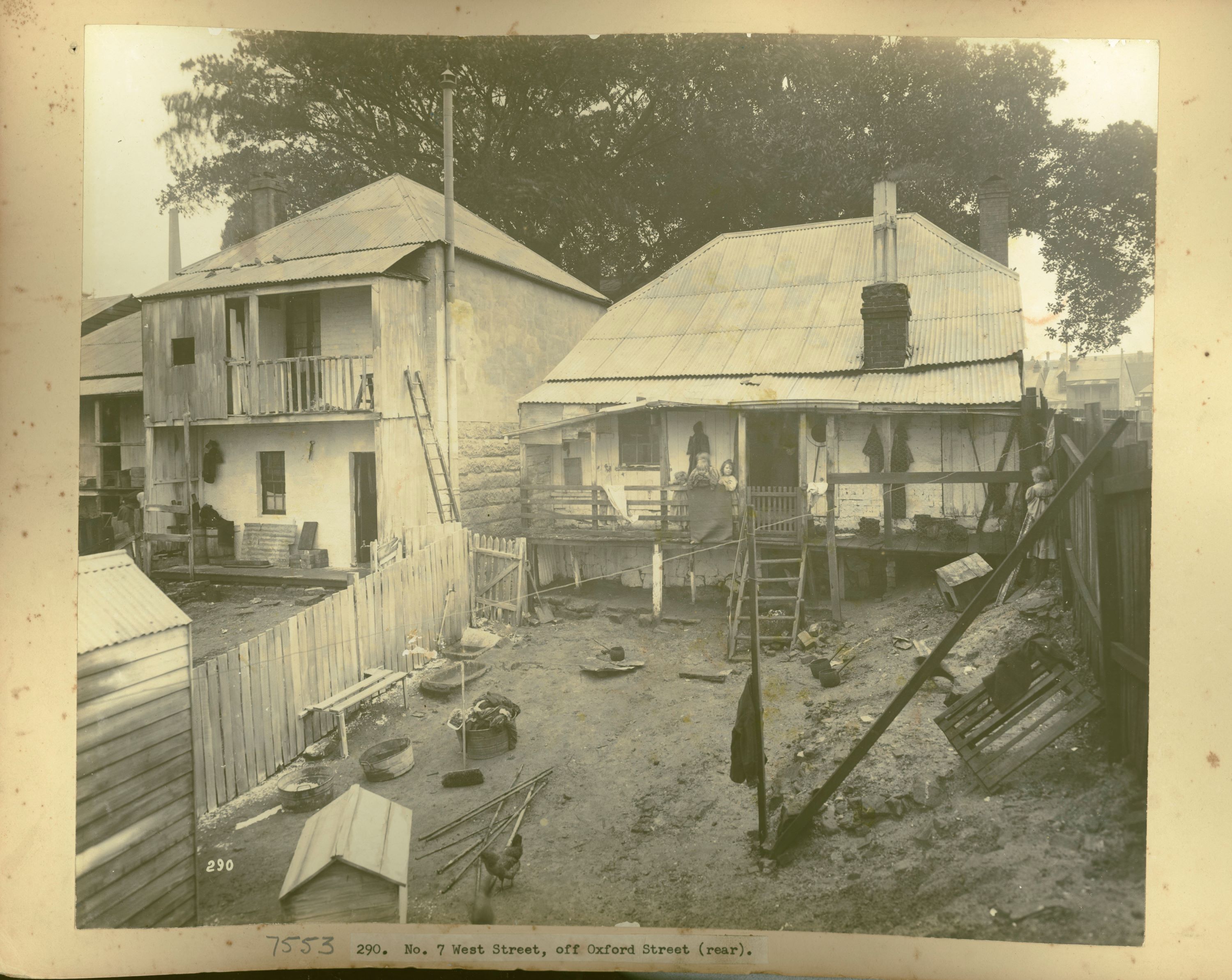 A group of children stand on the balcony of a wooden cottage looking over a muddy yard strewn with pots and cleaning tools. A crooked set of wooden steps leads to the balcony