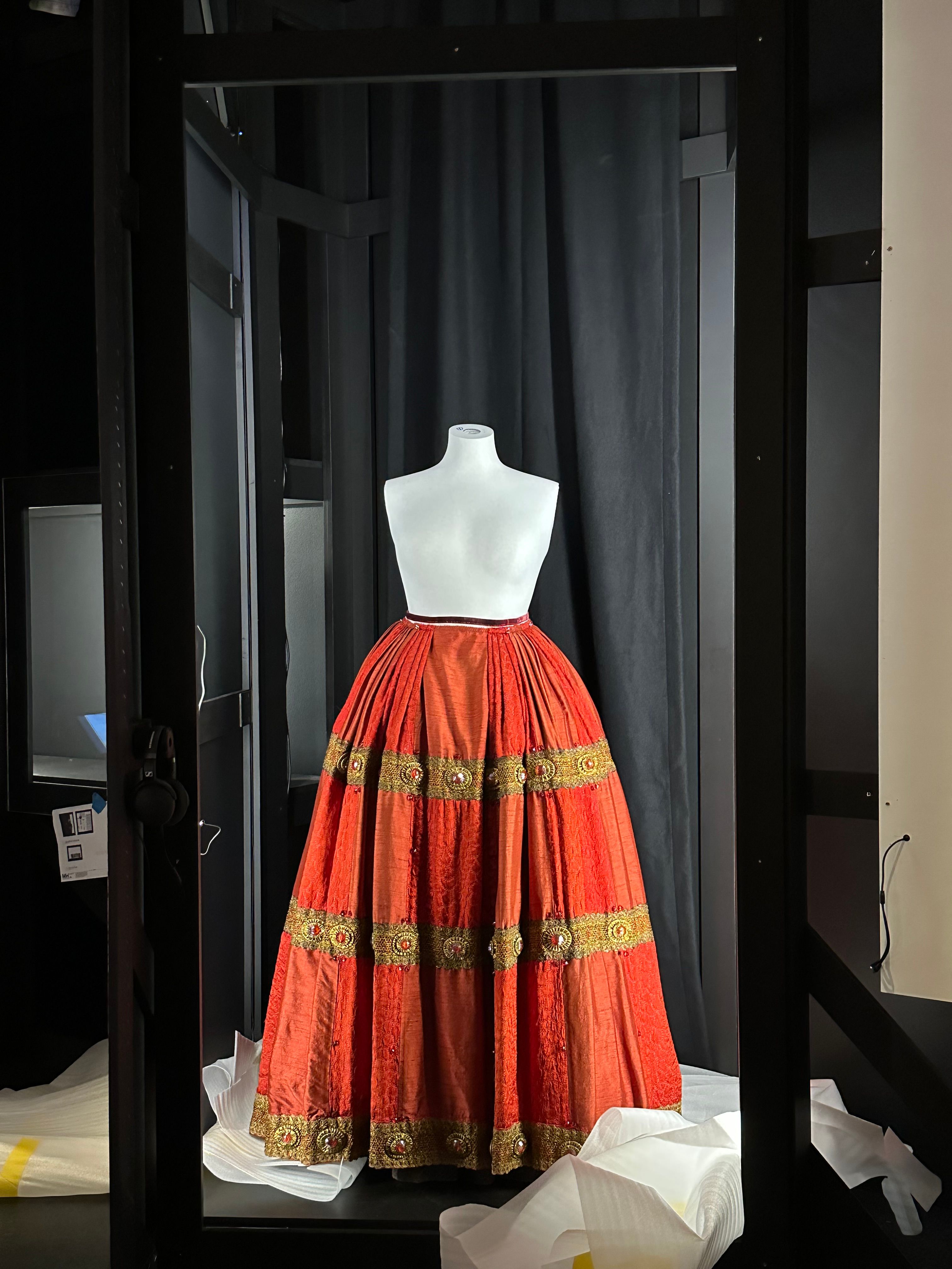 An elaborate full-bodied skirt in red material, with three horizontal bands of gold braid, has been placed on a plain white mannequin which does not yet have its arms or head attached.   