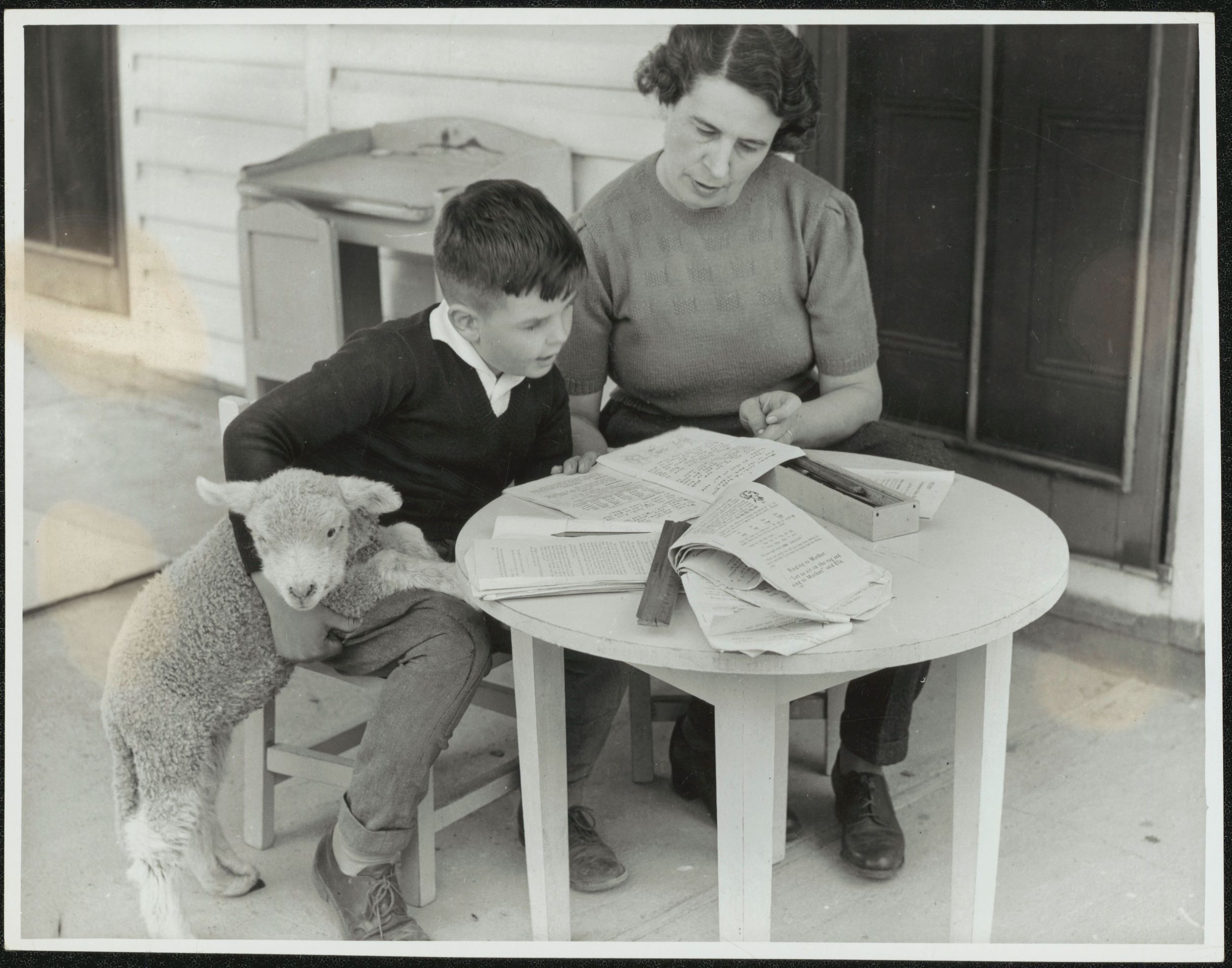 A boy and his mother sit at a table outside. A lamb rests its front legs on the boy's leg