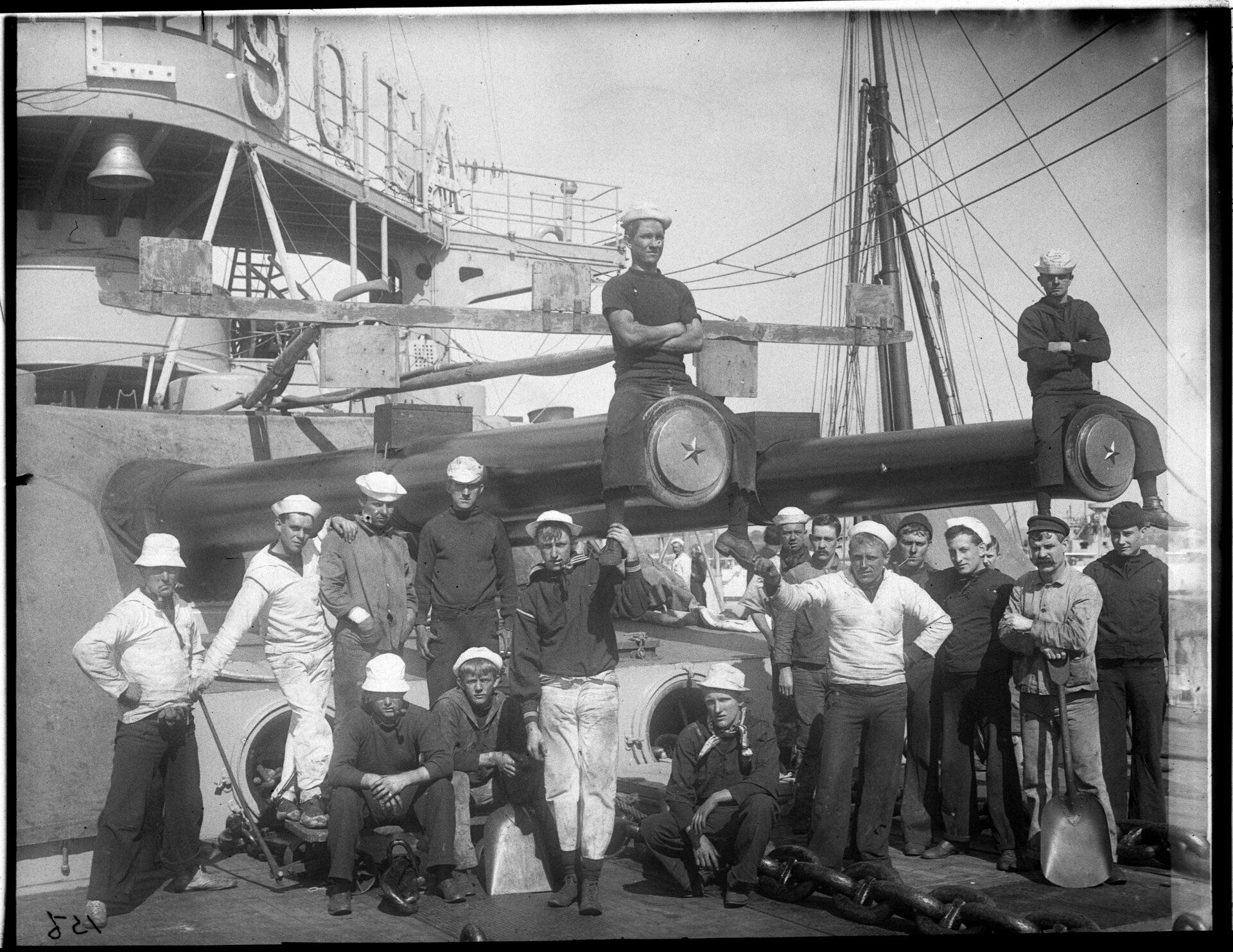  A group of sailors pose casually in front of a ship, leaning against and sitting on top of the cannons
