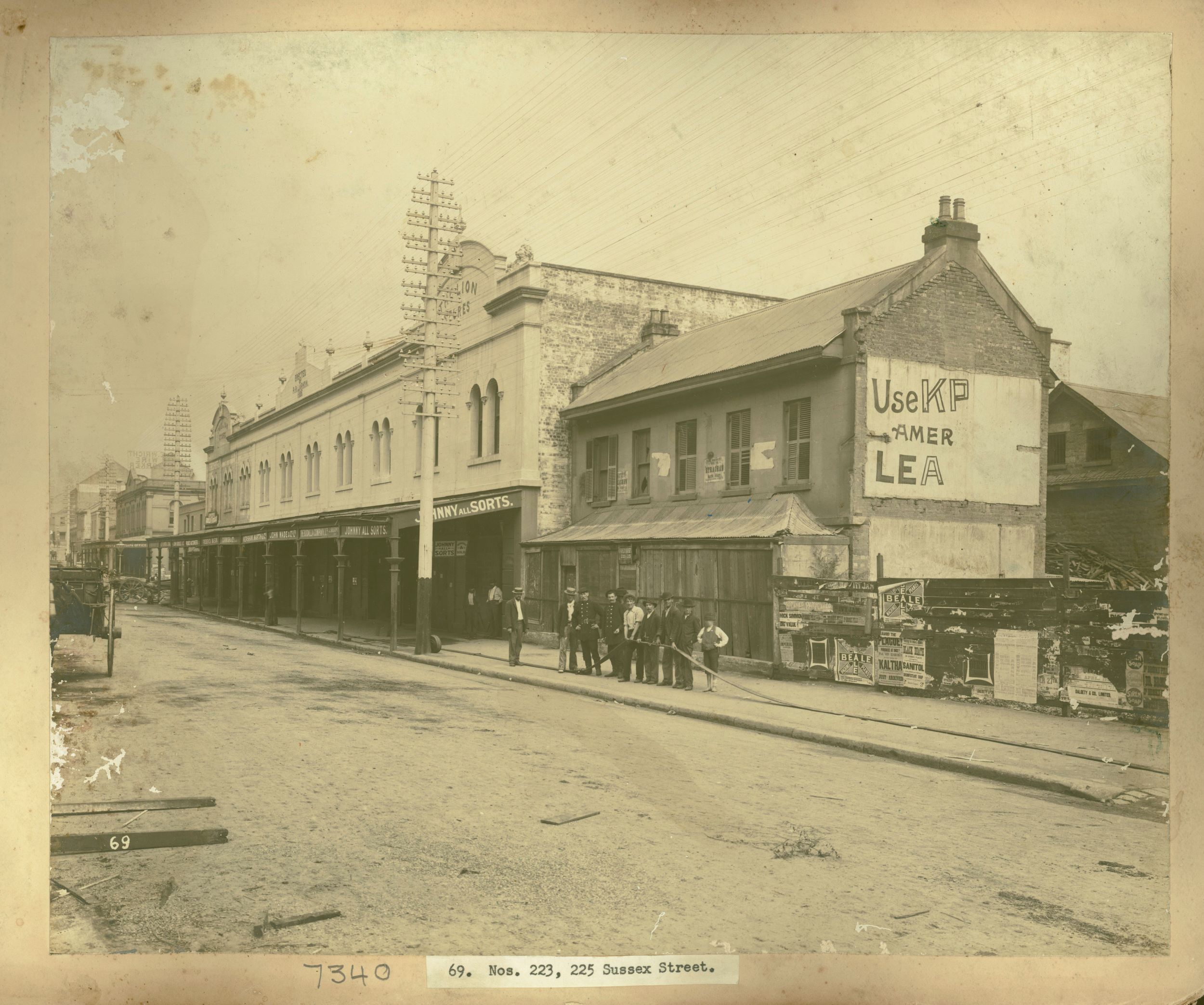 A Sydney street with a dirt road and a group of men stand in front of a row of shops with a hose