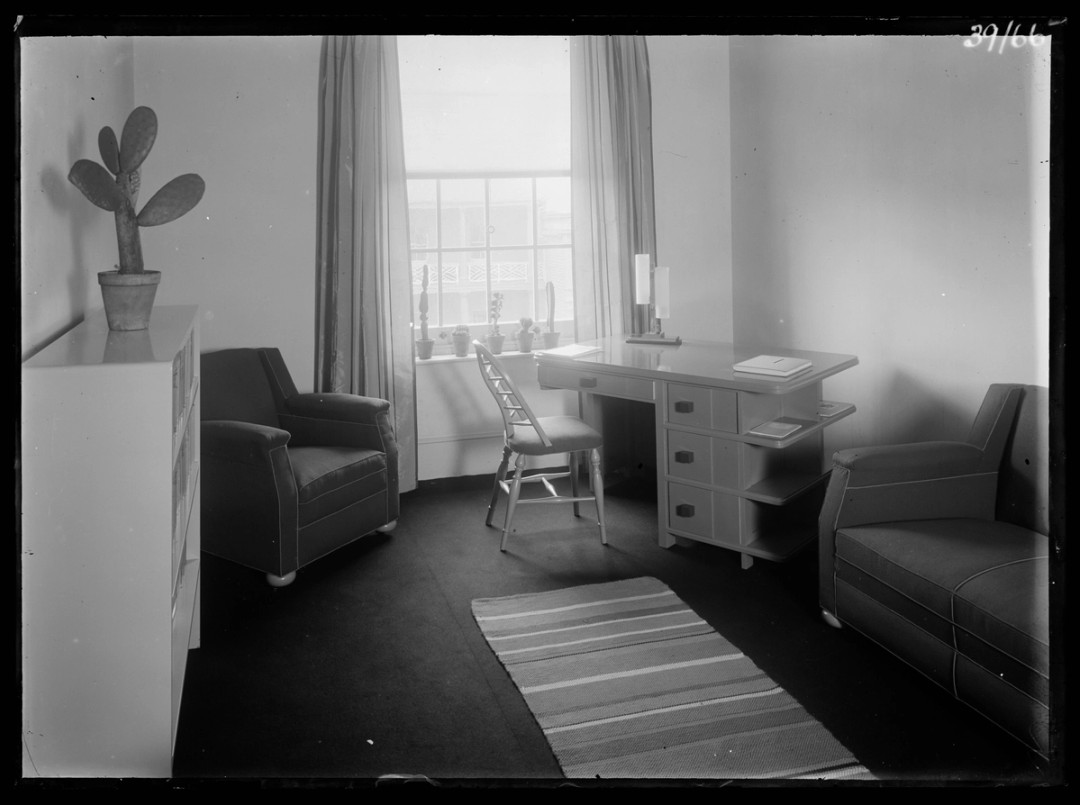 A study with desk, lounge and single armchair. A cactus plant sits atop shelving and a striped rug runs along the centre of the floor