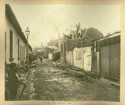 Onlookers watch a group of men demolishing the roof of a house. A pile of bricks has fallen into the muddy lane