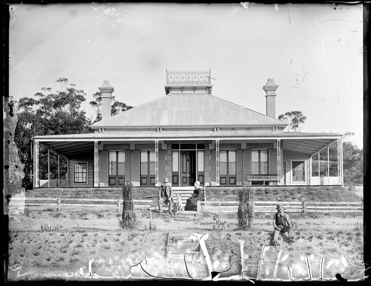 A man and woman sit at the foot of the stairs of a homestead with a wrap around verandah. A dog sits between them