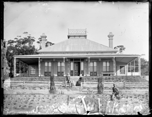 A man and woman sit at the foot of the stairs of a homestead with a wrap around verandah. A dog sits between them