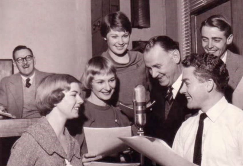 Group of people holding scripts standing around a microphone 