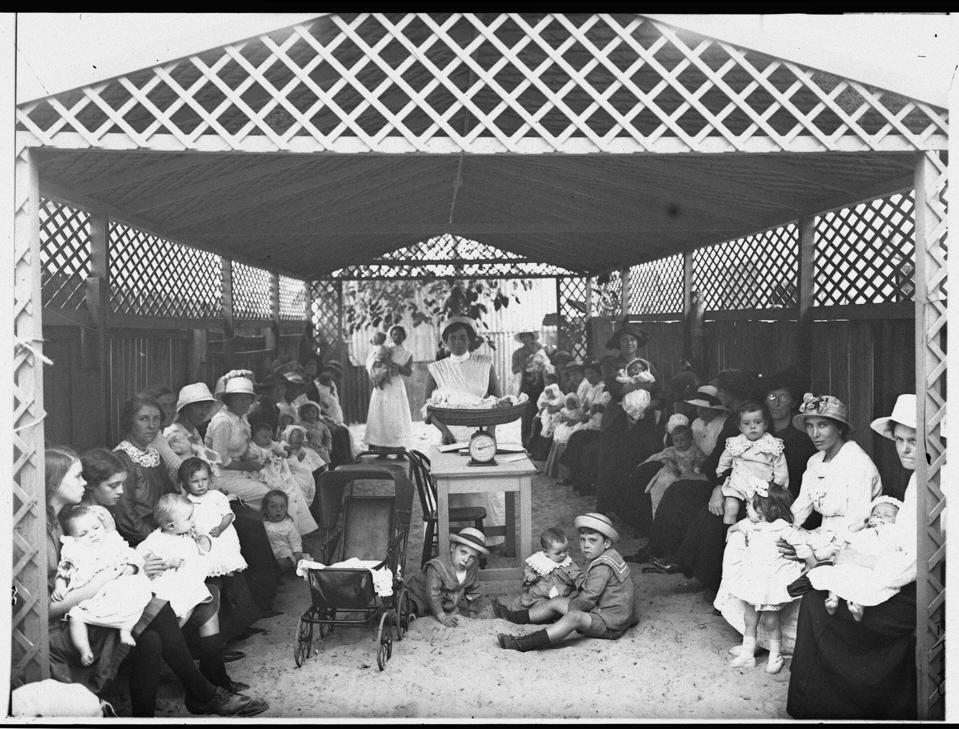 Mothers and their young children sit under an awning. In the centre a nurse stands at a table that holds a baby in a weighing machine