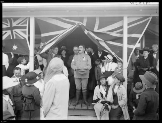 A soldier stands on a stage with the British and Australian flags decorating either side. A crowd is before him and several people stand behind him 