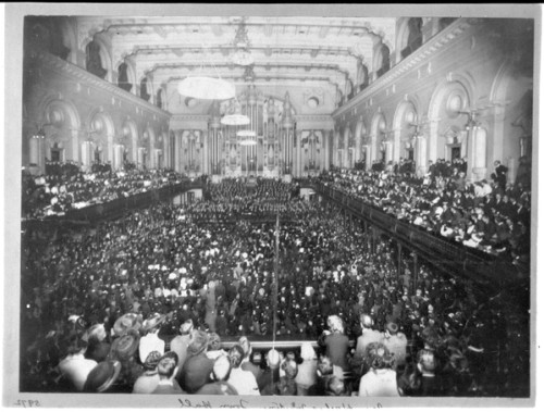 A hall is crowded with people sitting in rows down the centre of the floor and in balconies at either side