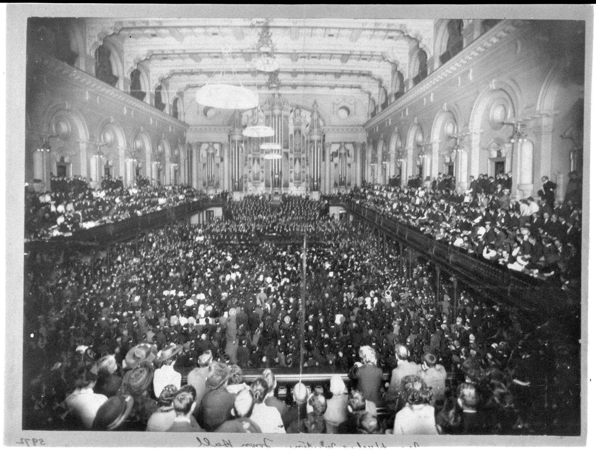 A hall is crowded with people sitting in rows down the centre of the floor and in balconies at either side