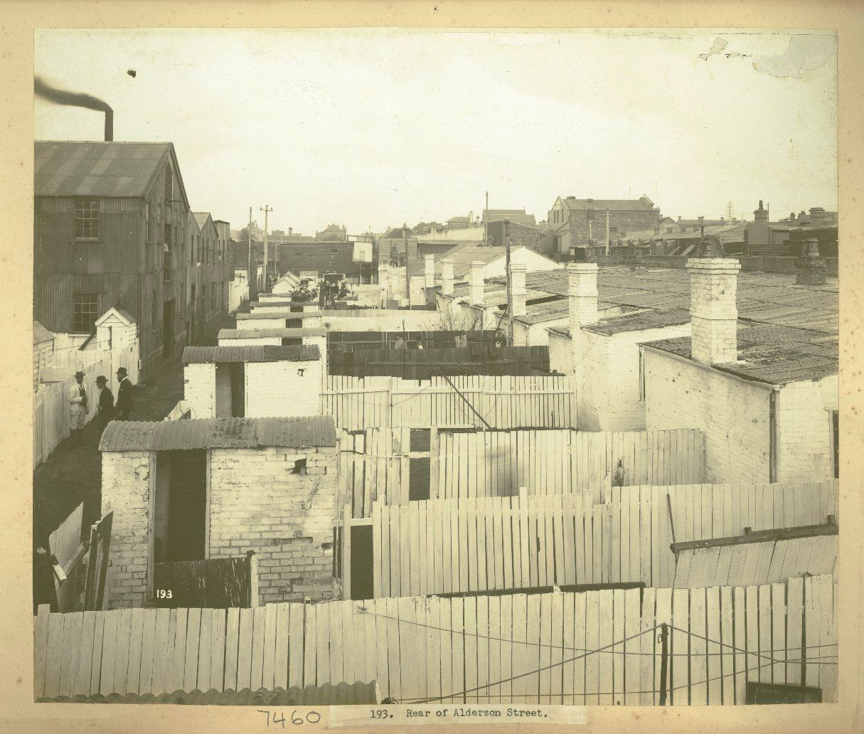 A long row of uniform backyards with white fences and outdoor lavatories. Three men stand in the laneway