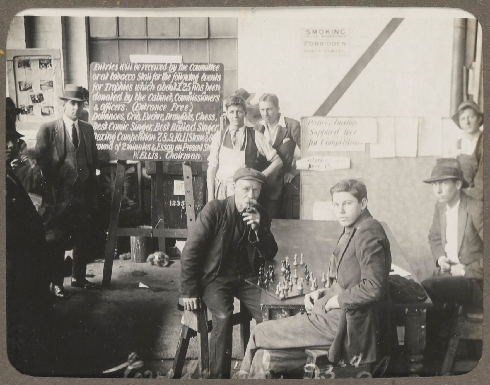 Two men sit at a table playing chess as others stand watching on