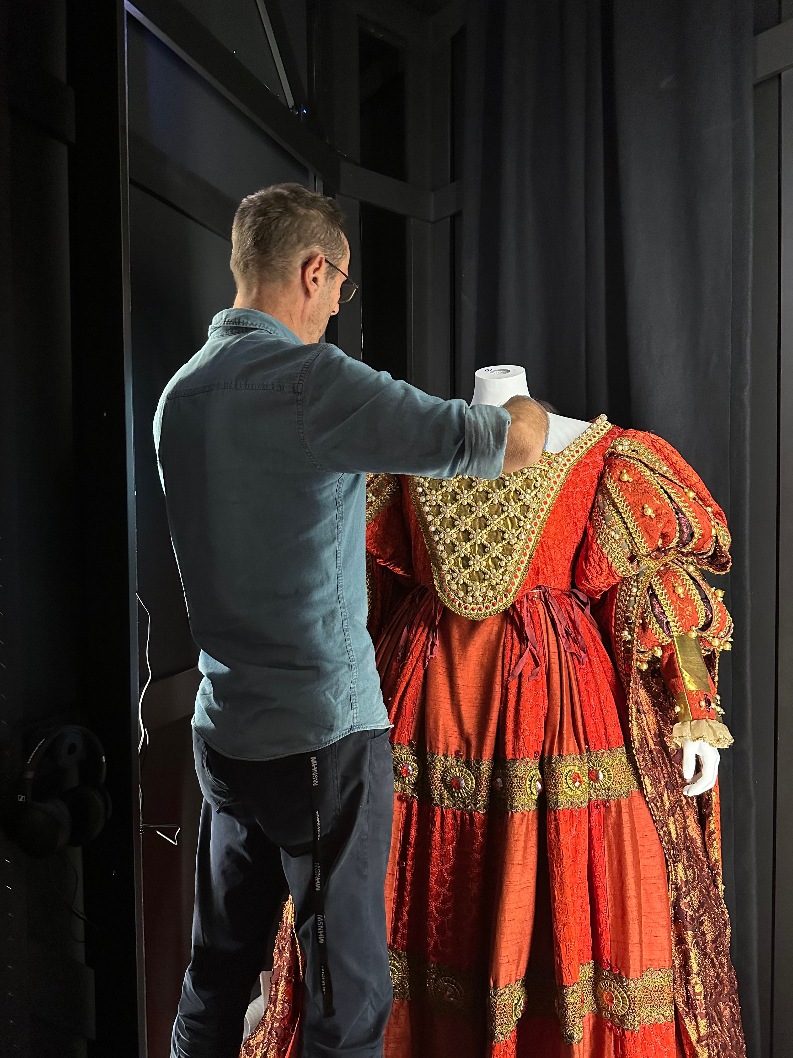 A man with his back to the viewer is adjusting the position of an elaborate stage costume on a mannequin which does not yet have its head attached. 