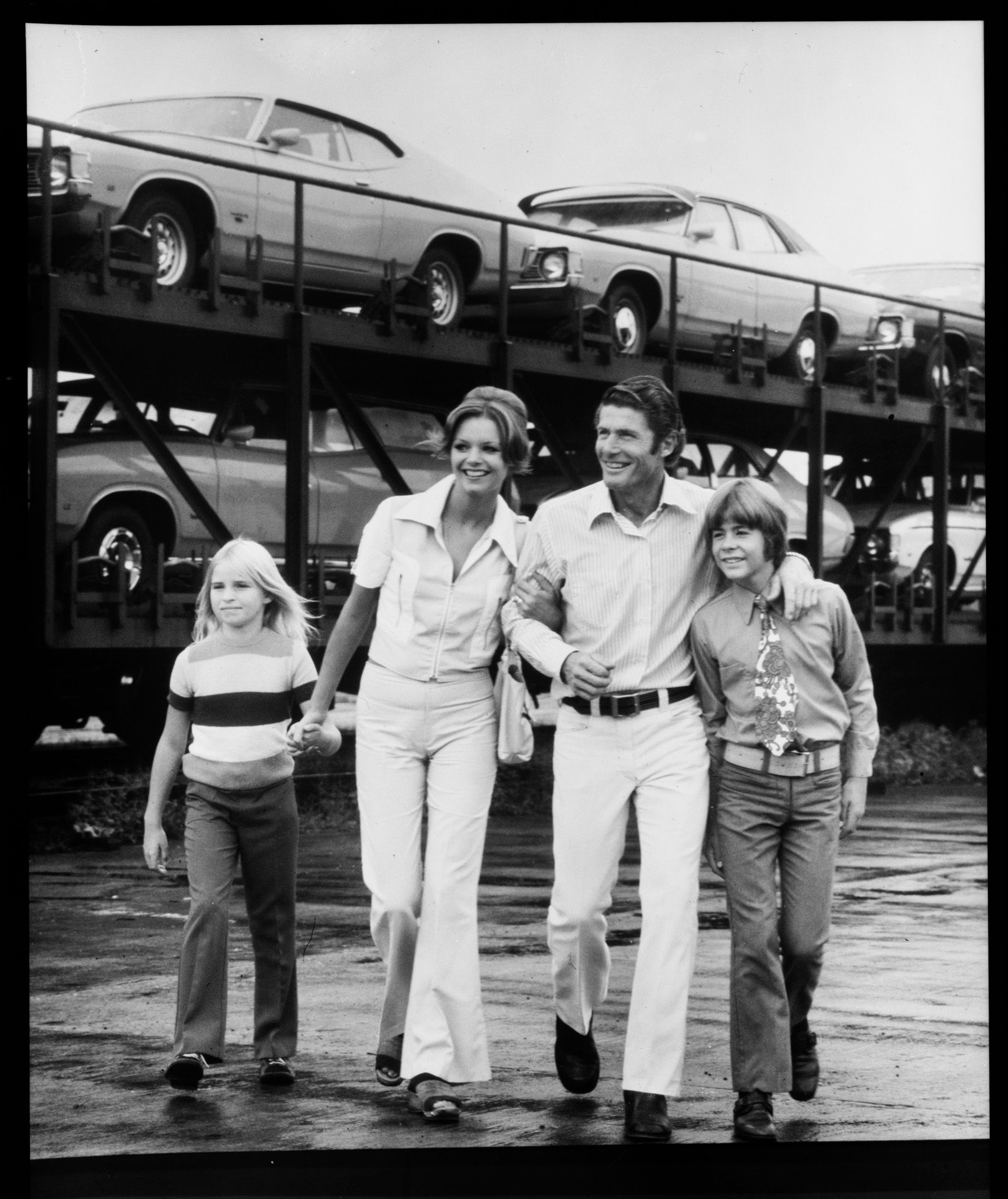 A family group stands in front of new cars on a trailer