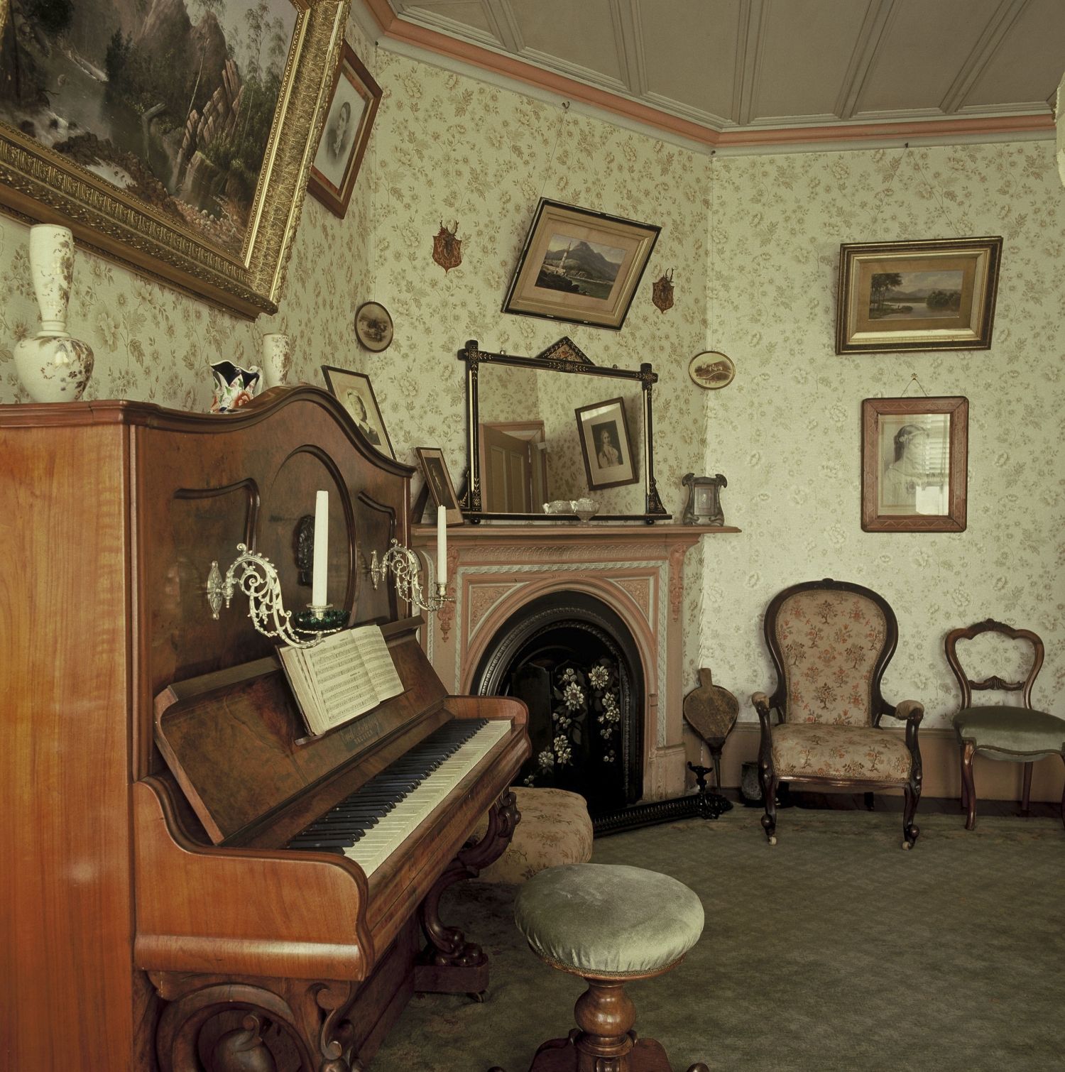 Colour photo of room with piano, fireplace, chairs, ornaments and frame pictures on wallpapered walls.