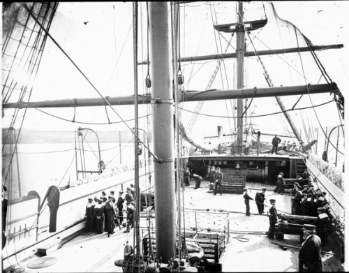 Boys in nautical clothing on top deck stand at the cannons lining the sides of the ship while instructors look on