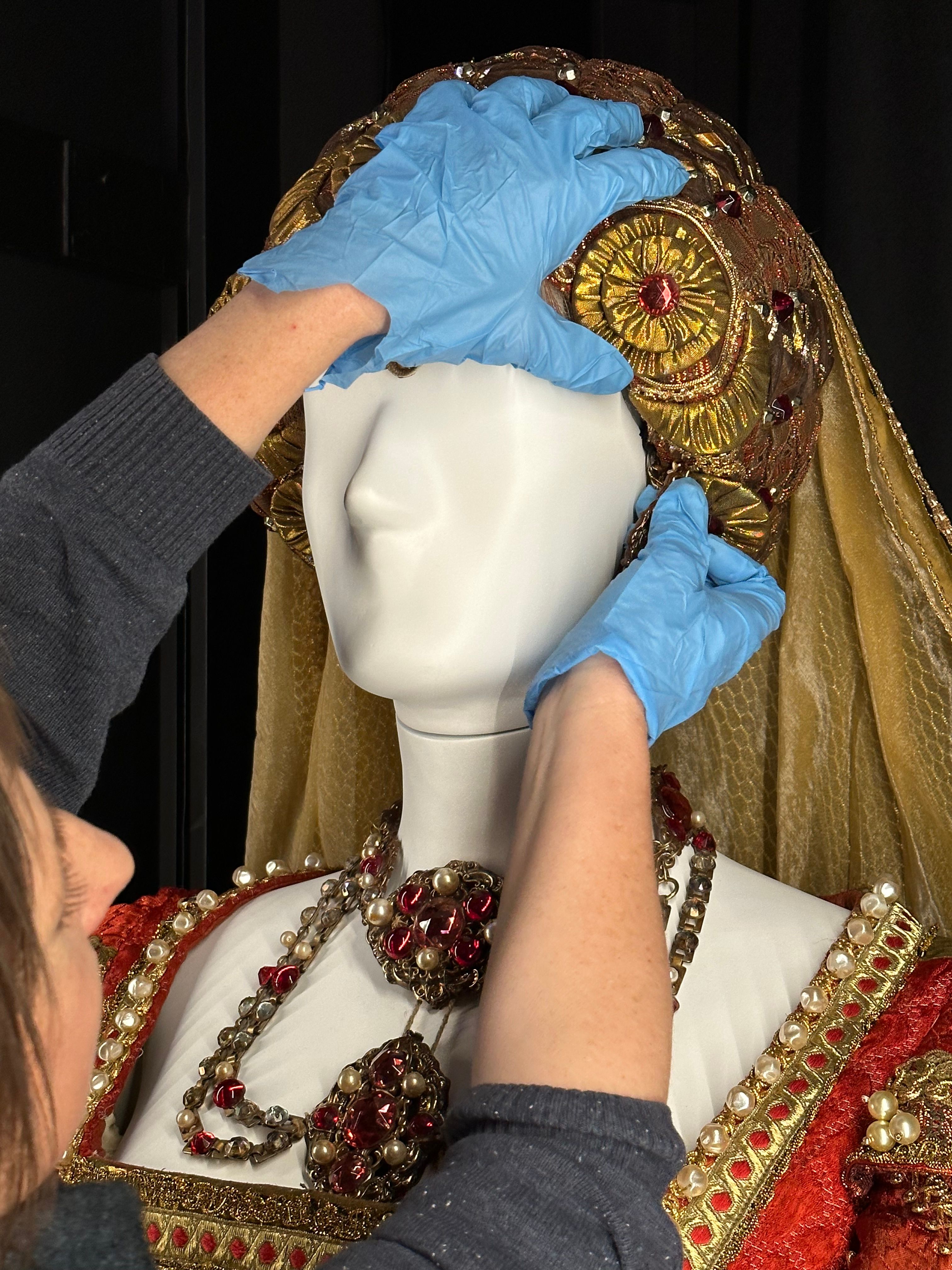 A woman wearing blue gloves is reaching up to secure a gold-coloured headdress on the head of a white-coloured mannequin, on which the shoulders of an elaborate bodice in red material with gold braid, and a necklace resembling gold set with rubies and pearls can also be seen. 