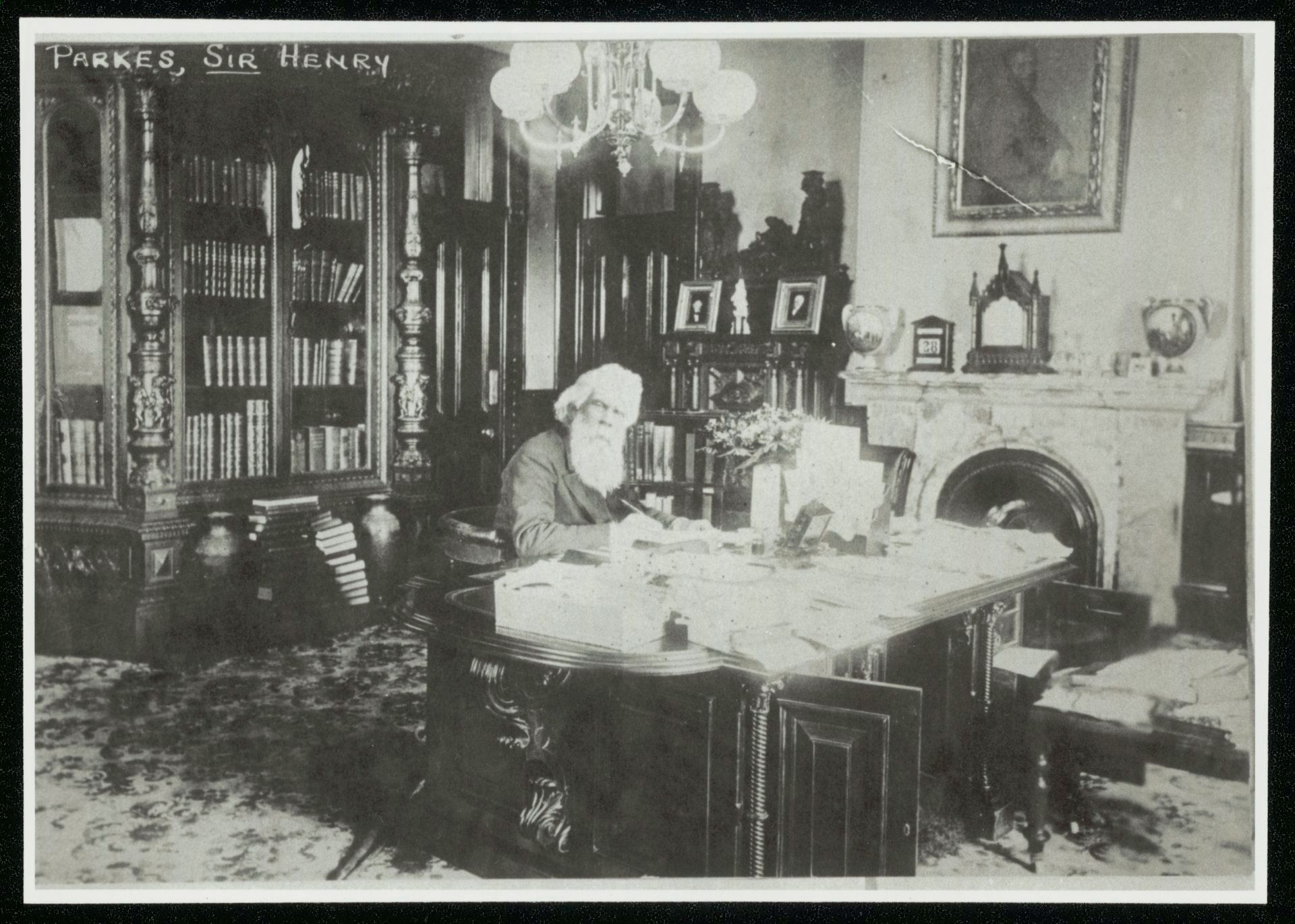 A man sits at a desk in a large office surrounded by bookshelves and a fireplace