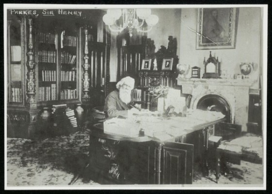 A man sits at a desk in a large office surrounded by bookshelves and a fireplace